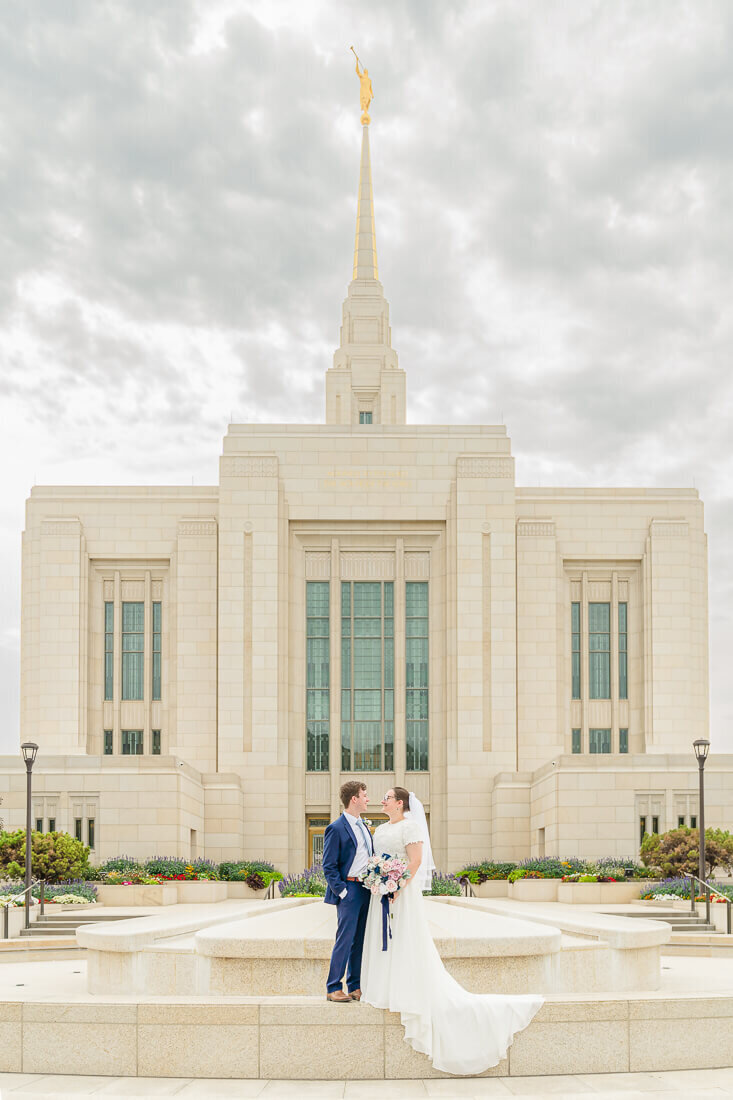 Bride and groom holding a bouquet of pink and white roses with a navy silk ribon standing at a fountain facing each other with a full view of the ogden temple