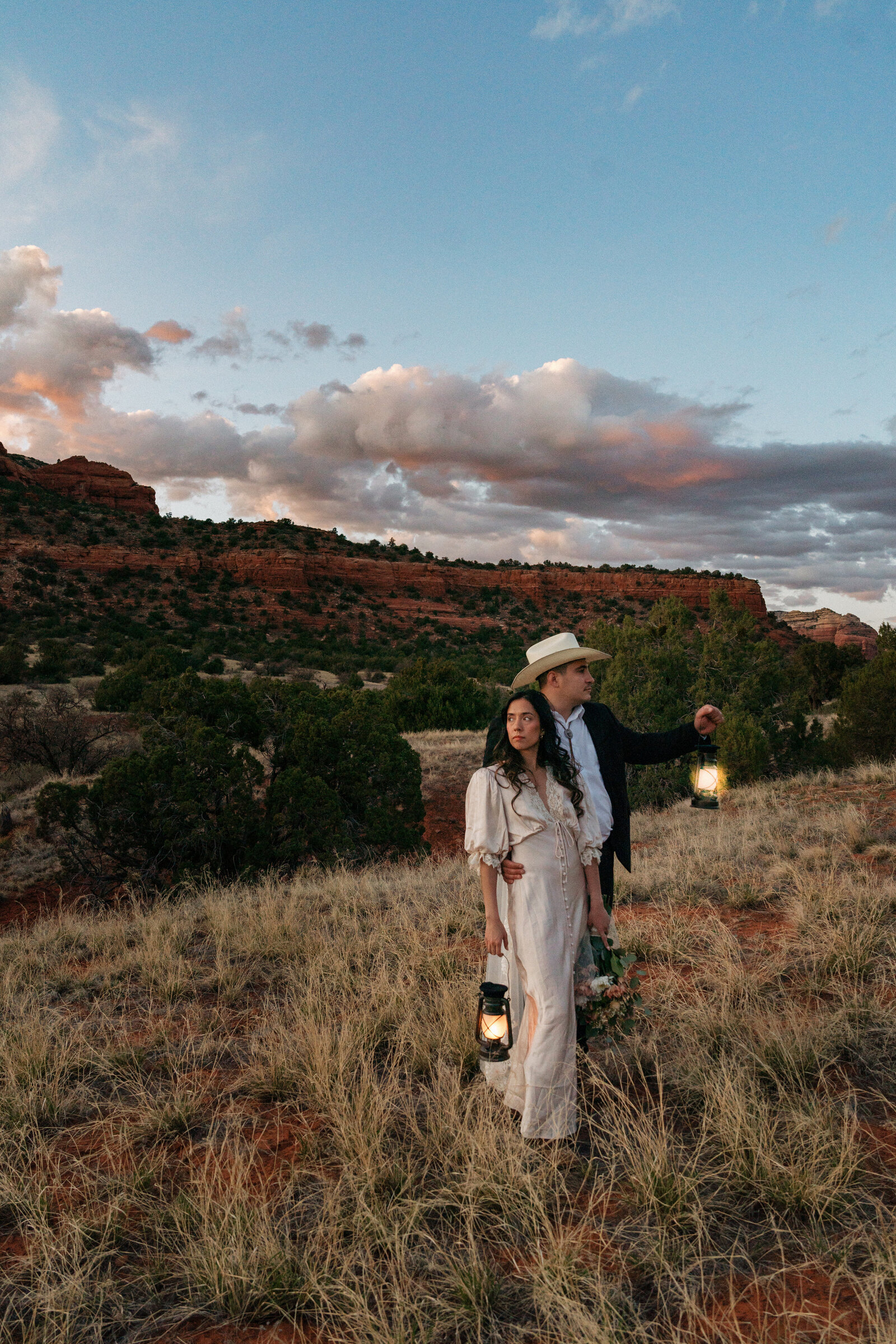 Bride and groom in western attire holding lanterns during their Bear Mountain elopement in Sedona, walking through desert grass at sunset with dramatic clouds overhead.