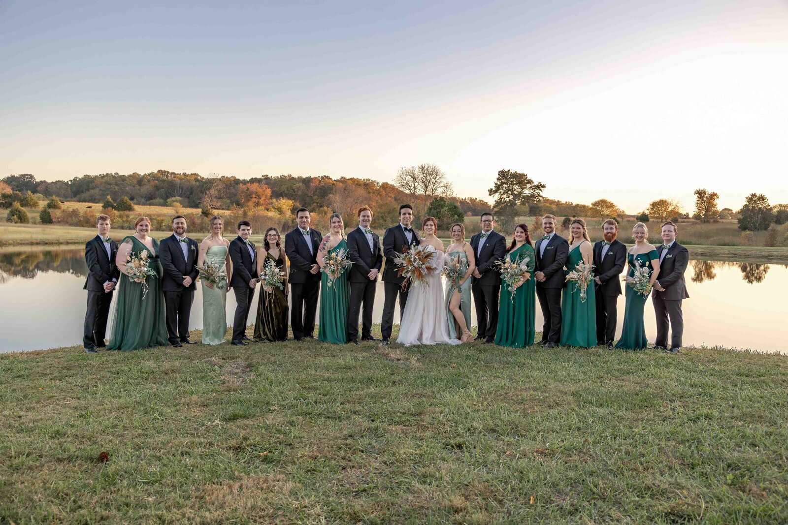 A large wedding party dressed in formal attire poses by a reflective pond at sunset during a Boone, NC wedding, blending rustic elegance with natural beauty and autumn colors.