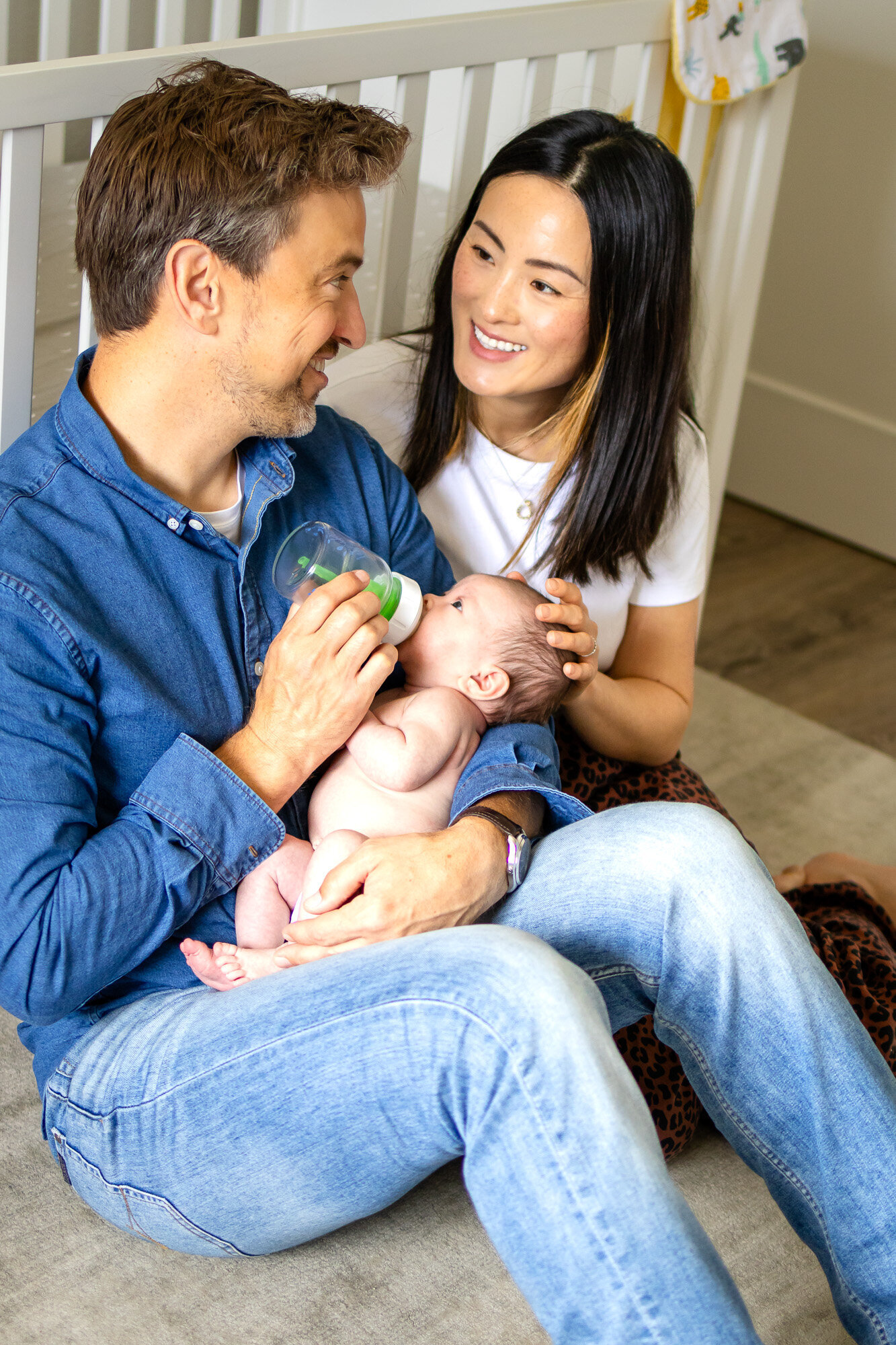 A dad feeds his baby while the mom smiles lovingly—an intimate in-home moment by Ellobelle Photography.