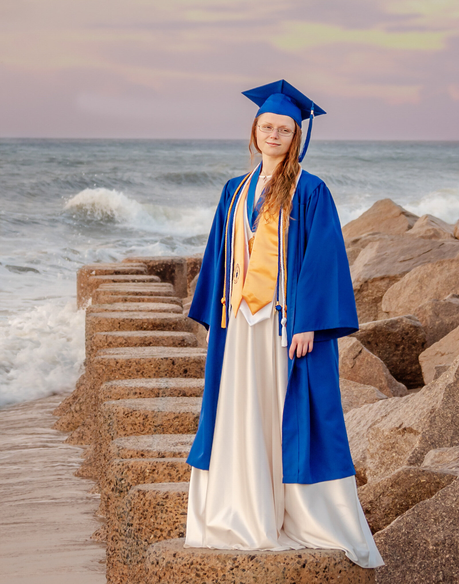 A college senior in a blue cap and gown standing on beach rocks with waves behind in Wilmington, North Carolina. This striking photo highlights the celebration of graduation, perfect for capturing memorable senior moments by the sea.