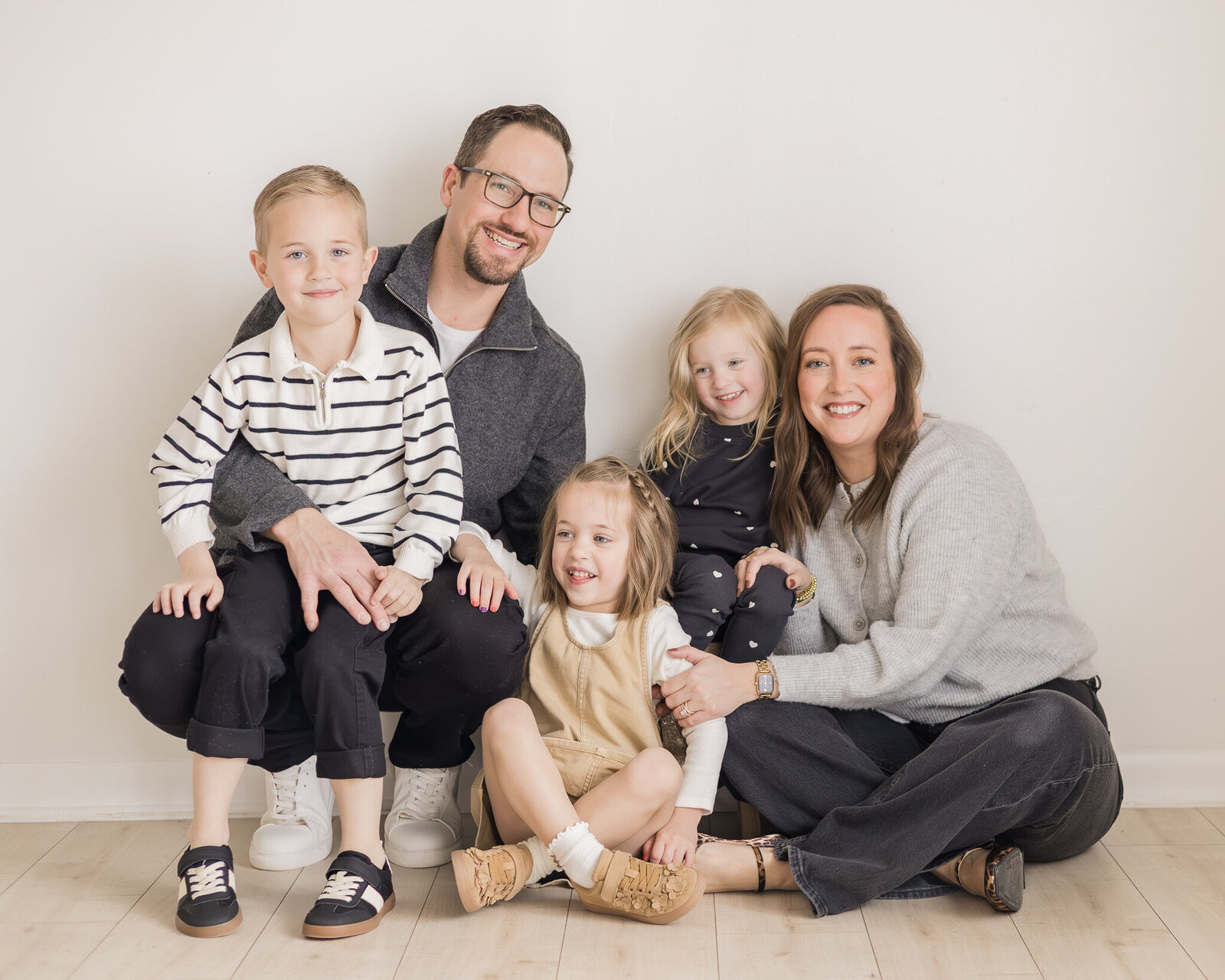 A family of 5 sitting on a light brown floor in a studio against a wall.  Dad is wearing a dark gray sweater over a white shirt with black pants, wearing glasses smiling at the camera.  Mom is wearing a light gray sweater, black pants, with dark brown hair smiling at the camera.  Young son has a white and blank striped shirt, black pants, and smiling at the camera.  Youngest daughter has blonde hair wearing a dark blue and dotted shirt and pants, smiling away from the camera.  Other daughter is wearing a light tan dress with a long sleeve white shirt, tan shoes, and smiling away from the camera