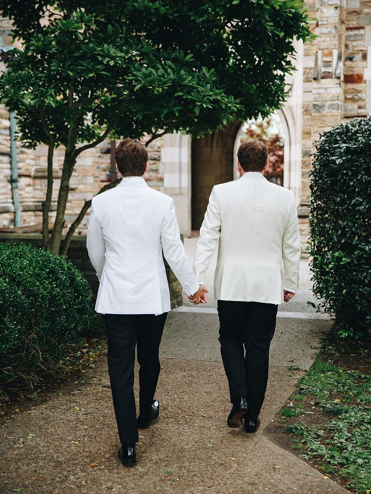 Two grooms wearing tuxedos with white jackets walk away hand in hand