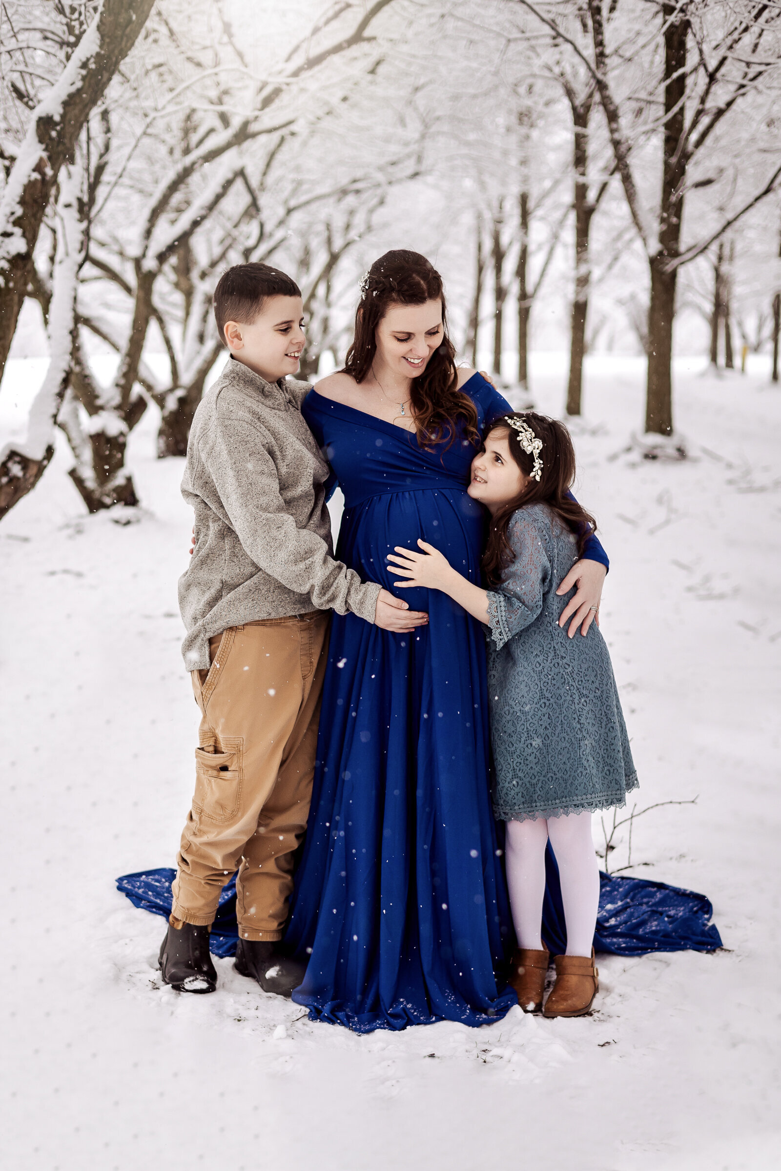 Pregnant mother in a blue gown  snuggles all three of her children on a snowy day