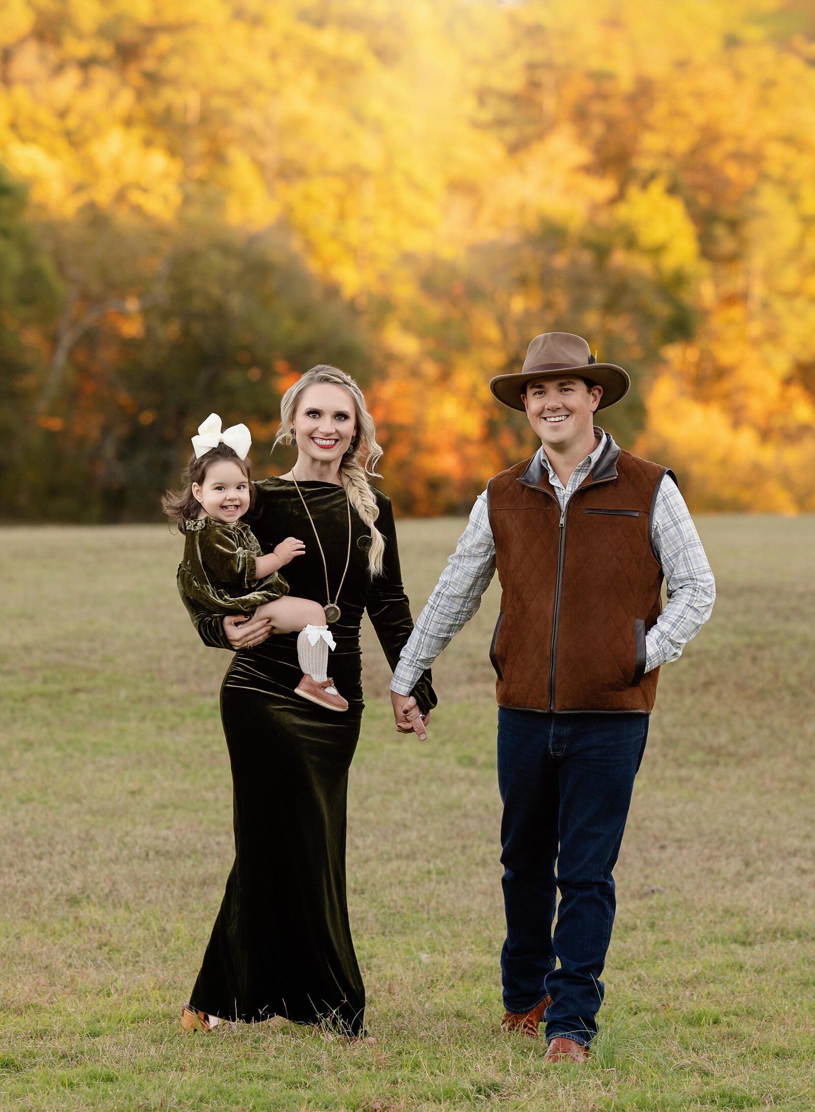 Family of 3 walking in open field with fall colors and smiling at camera.