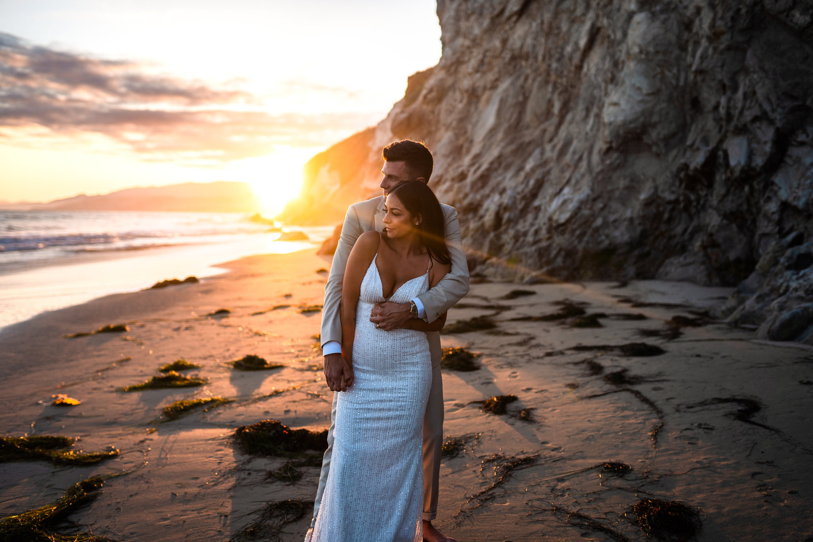 Couple in wedding attire, holding each other, and looking at towards the ocean during sunset.
