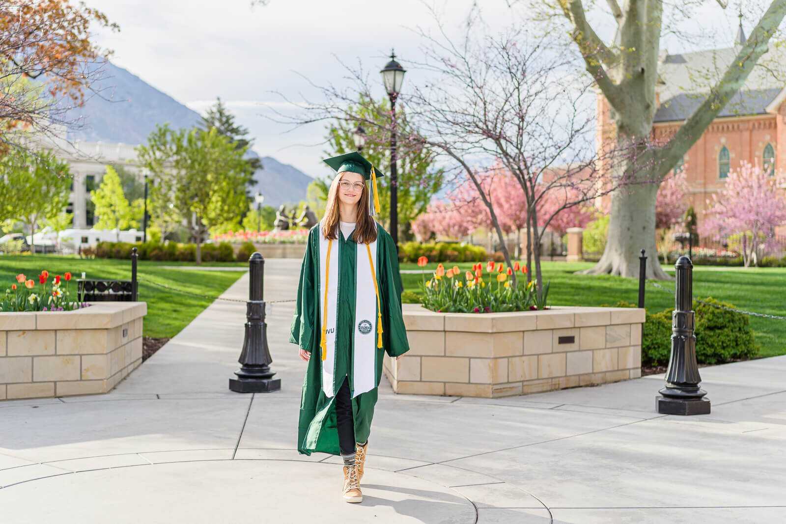 Salt Lake City senior photography of a high school senior girl wearing royal green colored graduation cap and gown walking in downtown Provo with blossoming trees in the distance