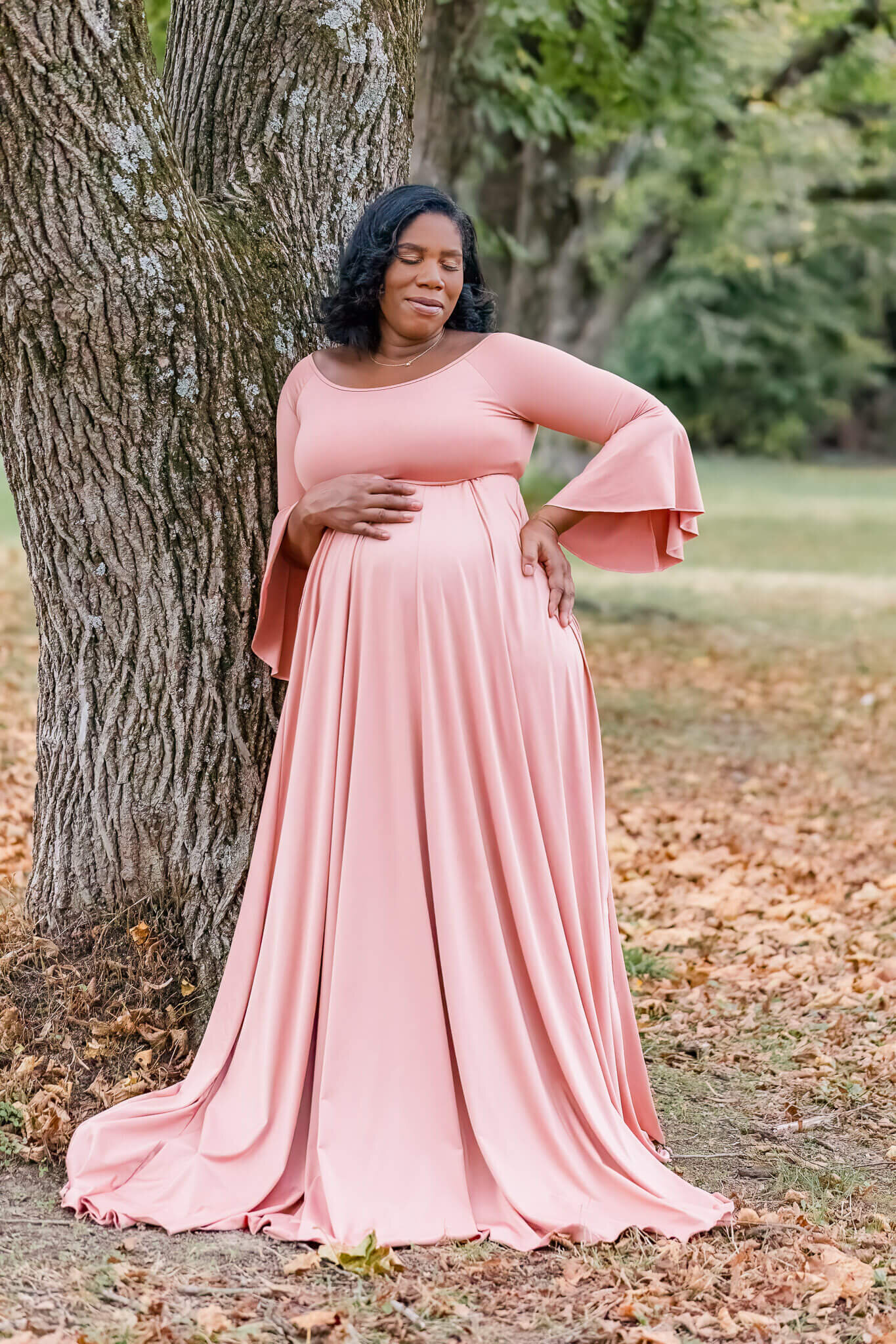 A pregnant woman embracing her belly and leaning against a tree at a park.