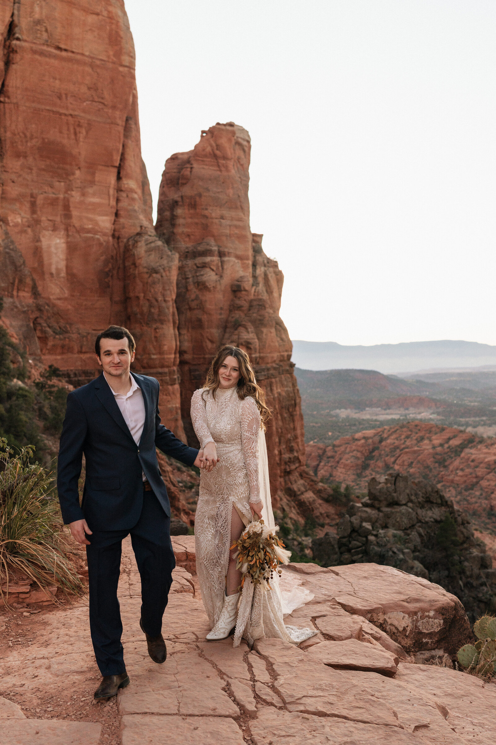 Bride in a lace dress and white boots holding a bouquet while walking hand-in-hand with the groom on the red rock cliffs of Cathedral Rock in Sedona, Arizona, surrounded by stunning desert views.