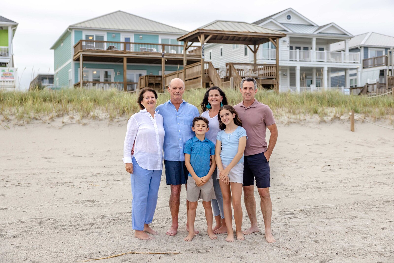 A family stands on the sandy shore, with beach houses as a backdrop. Dressed in blue and white, they capture a joyful seaside moment, perfect for artistic family photography in Wilmington, NC, highlighting family unity and coastal charm.