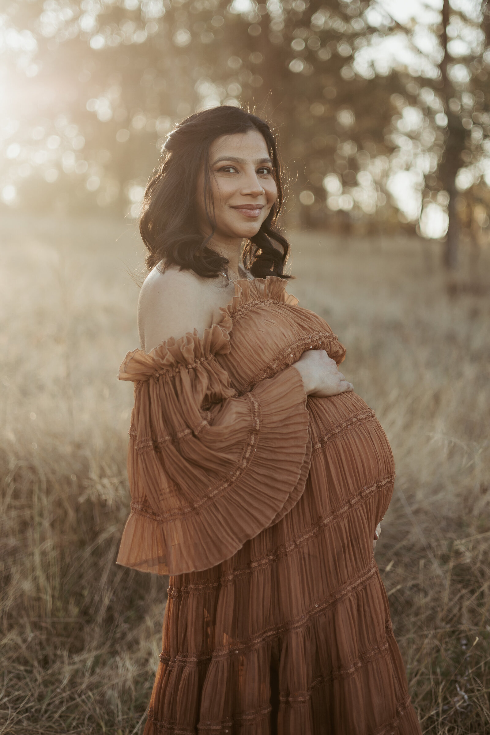 Glowing pregnant mama wearing a long brown dress looking directly at the camera