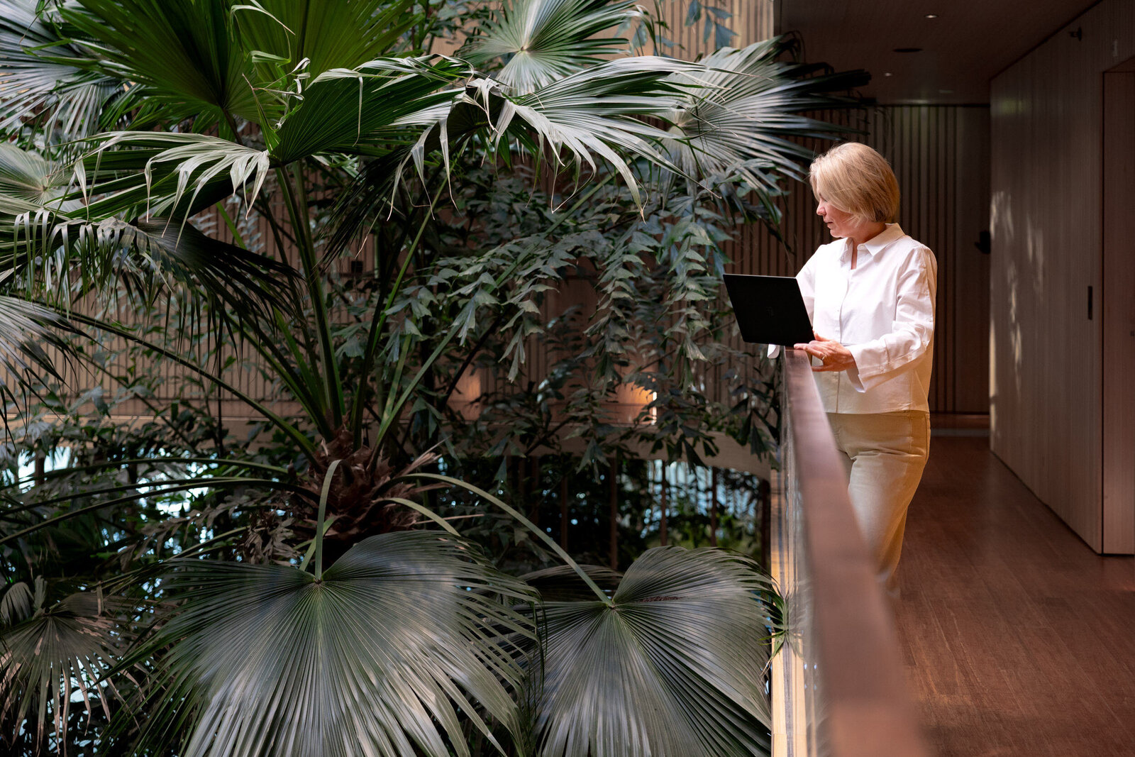 Burn-out coach Agnes Tuin staat met laptop in de gang van een groen werkomgeving met palmen