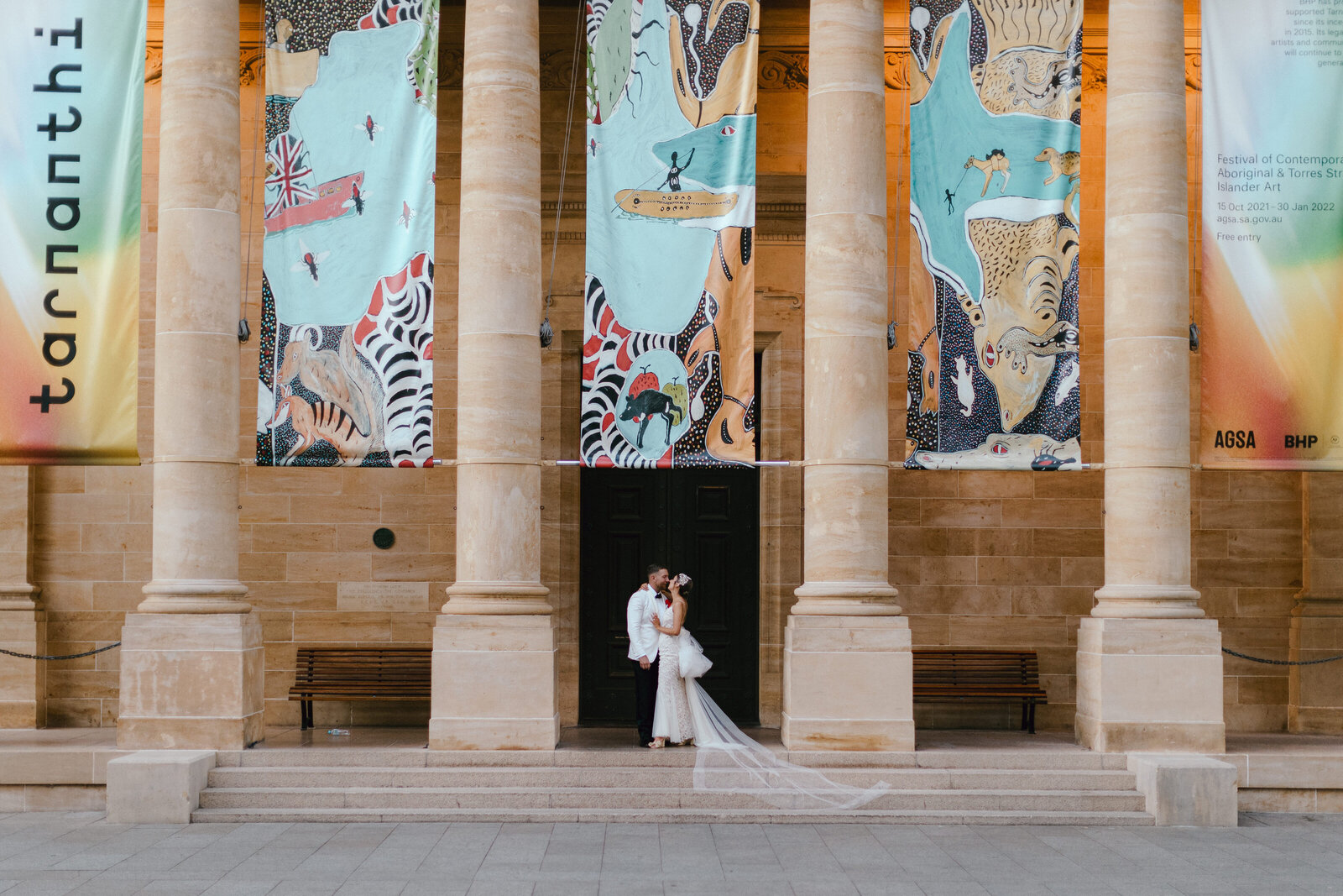 The façade of the Art Gallery of South Australia, tall limestone columns with a newlywed couple kissing in the centre.