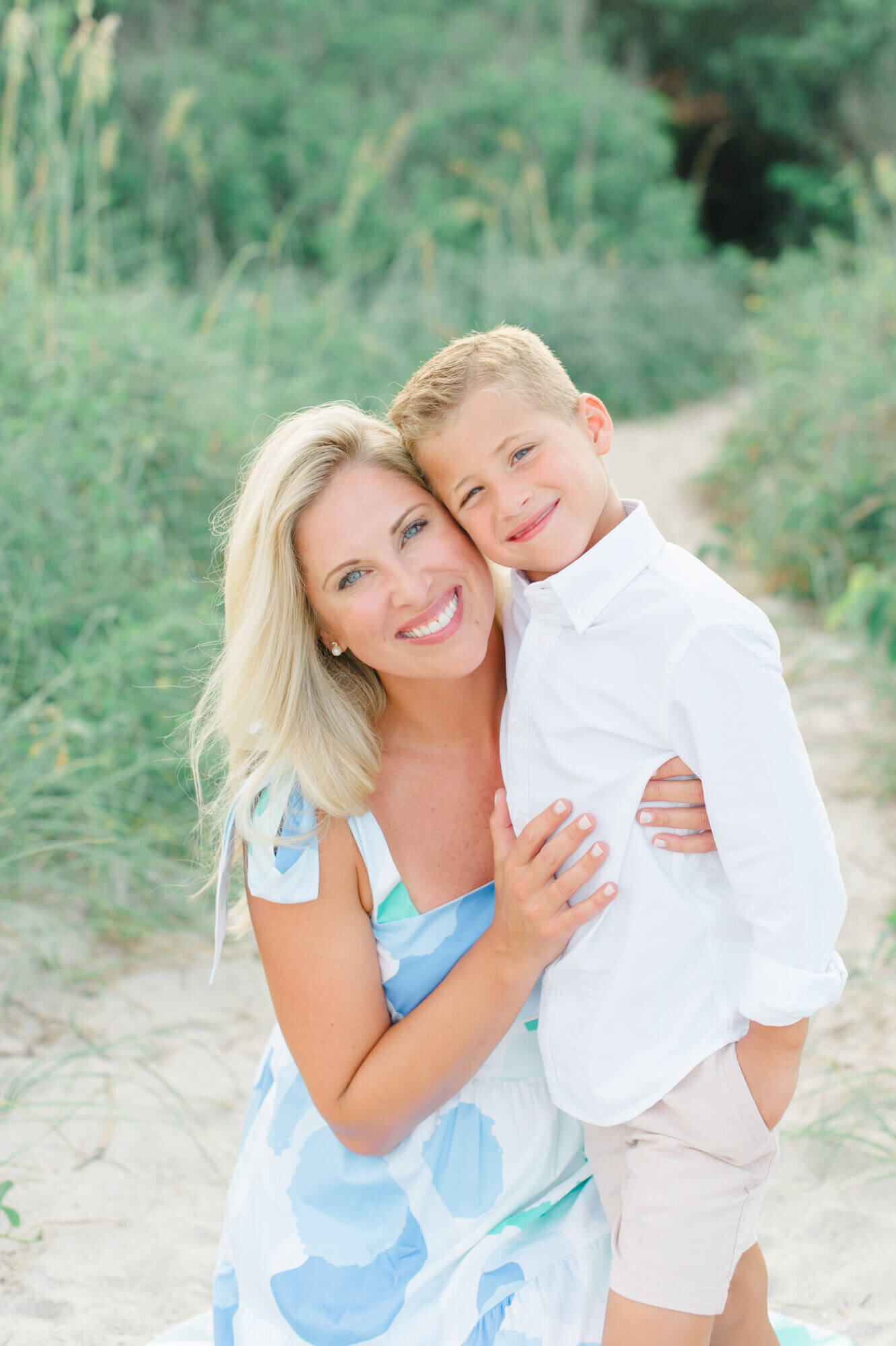 Mom holding her son near the beach dunes and greenery while smiling at the camera during their family beach photoshoot