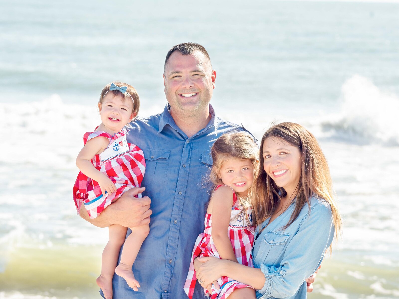  A joyful family poses together at Wrightsville Beach, showcasing two young children in matching red and white outfits, held lovingly by their parents under the bright sun. The ocean waves provide a serene backdrop, capturing the essence of a perfect family vacation. Ideal for those seeking an affordable family photography session in Wilmington, NC.