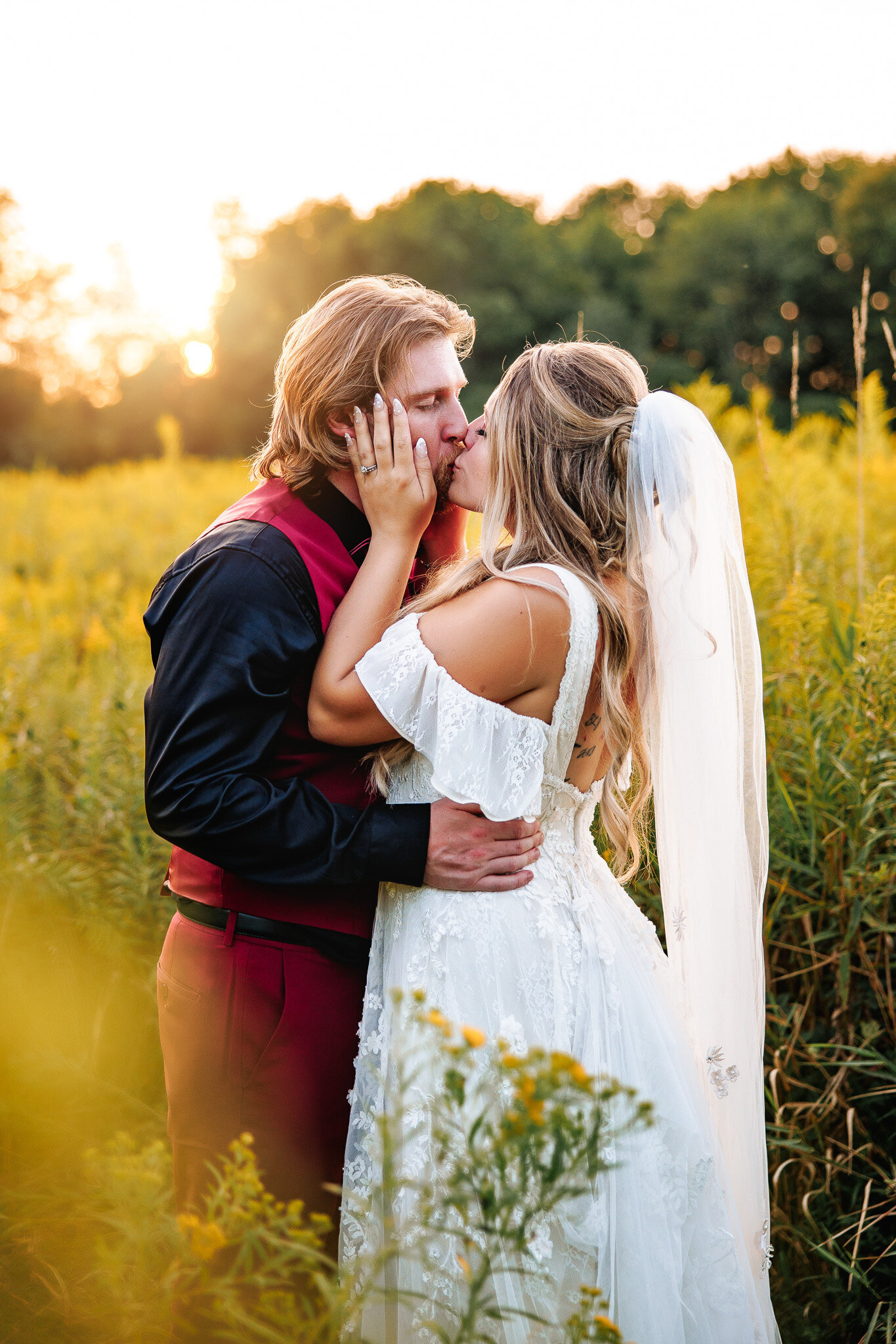 With her and on his cheek and his arm around her waist, a bride and groom sharer a kiss amongst a field of tall grasses