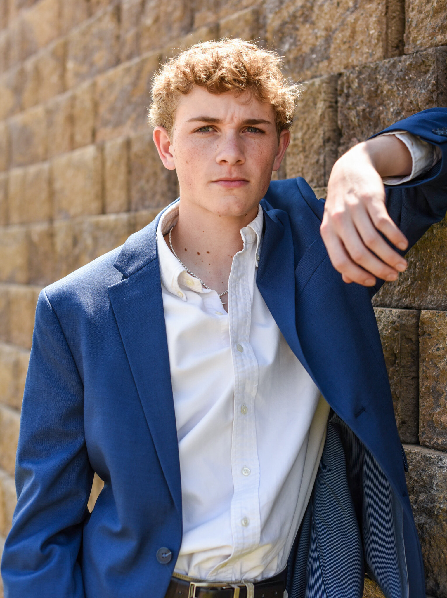 a senior dressed in a blue suit jacket leaning against a brick wall as he poses for his graduation photos photographed by Millz Photography in Greenville, SC