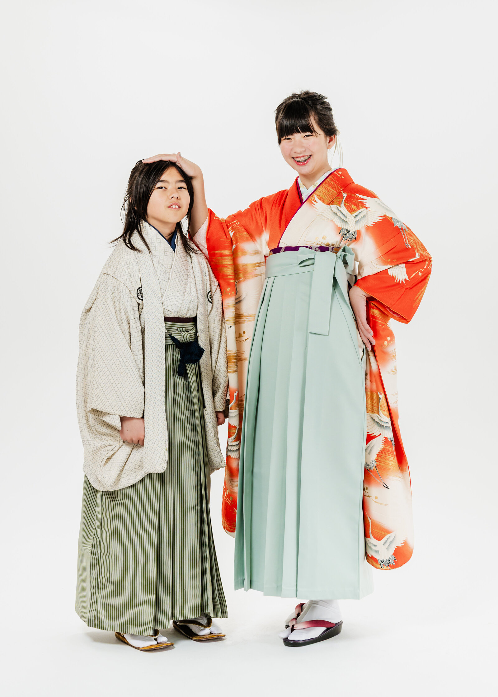Siblings in graduation kimono—girl in vibrant orange with green hakama playfully patting younger brother’s head | 卒業式 兄妹 姉 オレンジ着物 グリーン袴 弟 頭に手