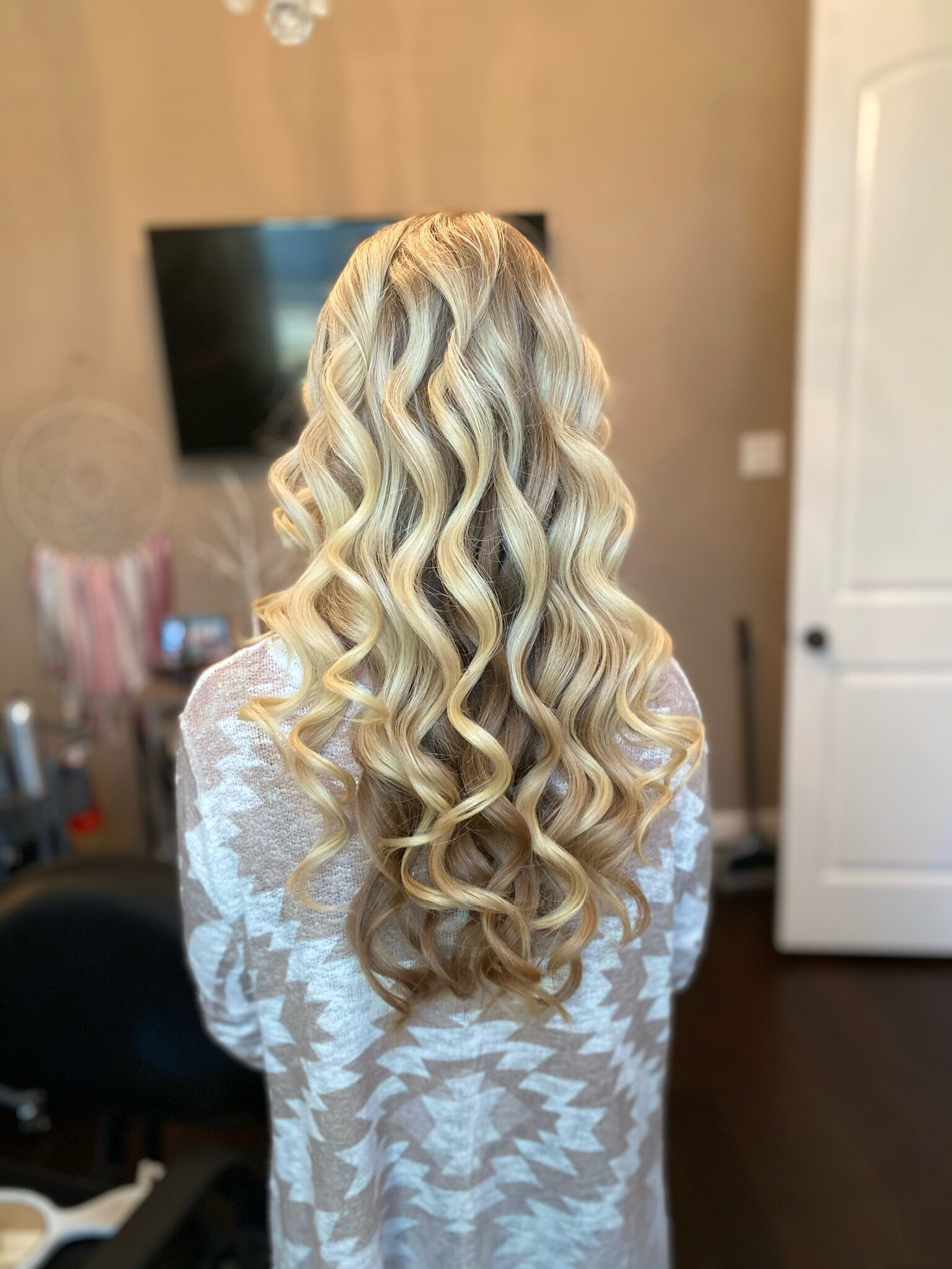 A woman with long, curly blonde hair faces away in a cozy room with soft lighting, perhaps savoring her upcoming bridal beauty experience in Texas.