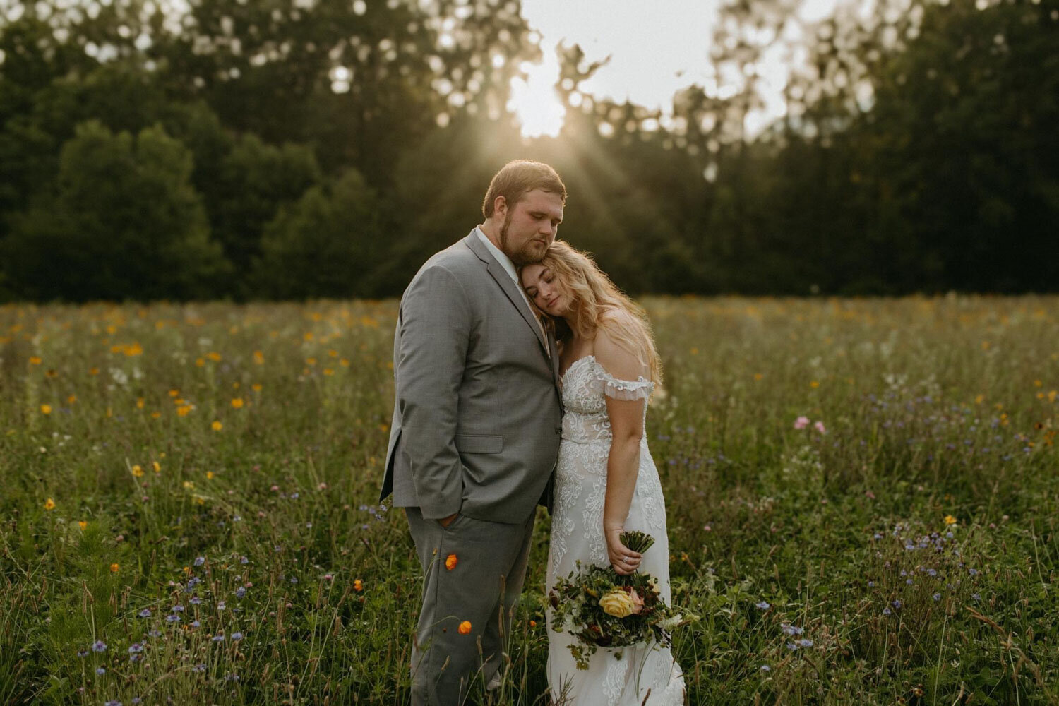 Bride resting her head on groom’s shoulder in a sunlit wildflower field