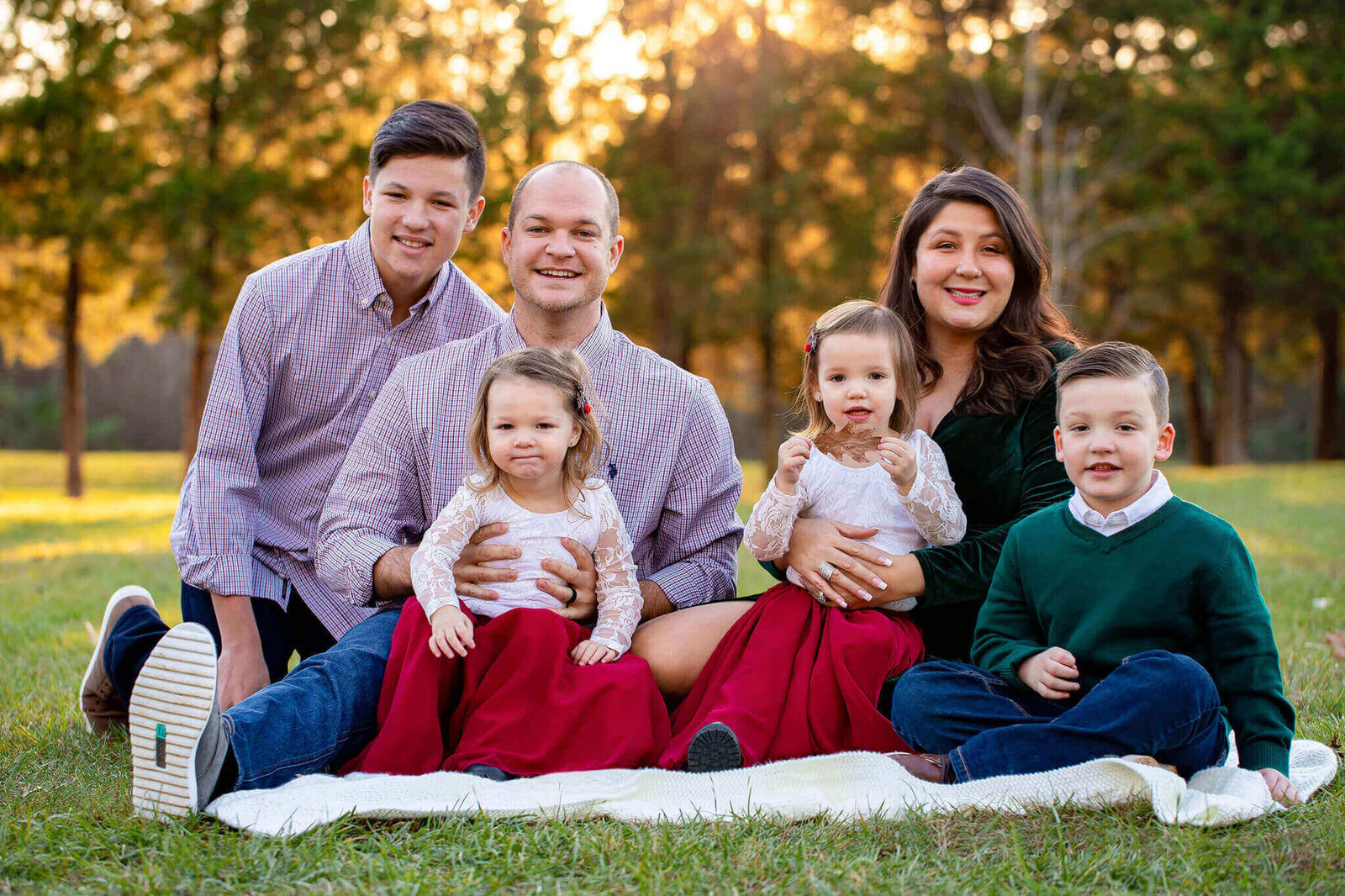 A family of six sitting on a blanket at a park at sunset.