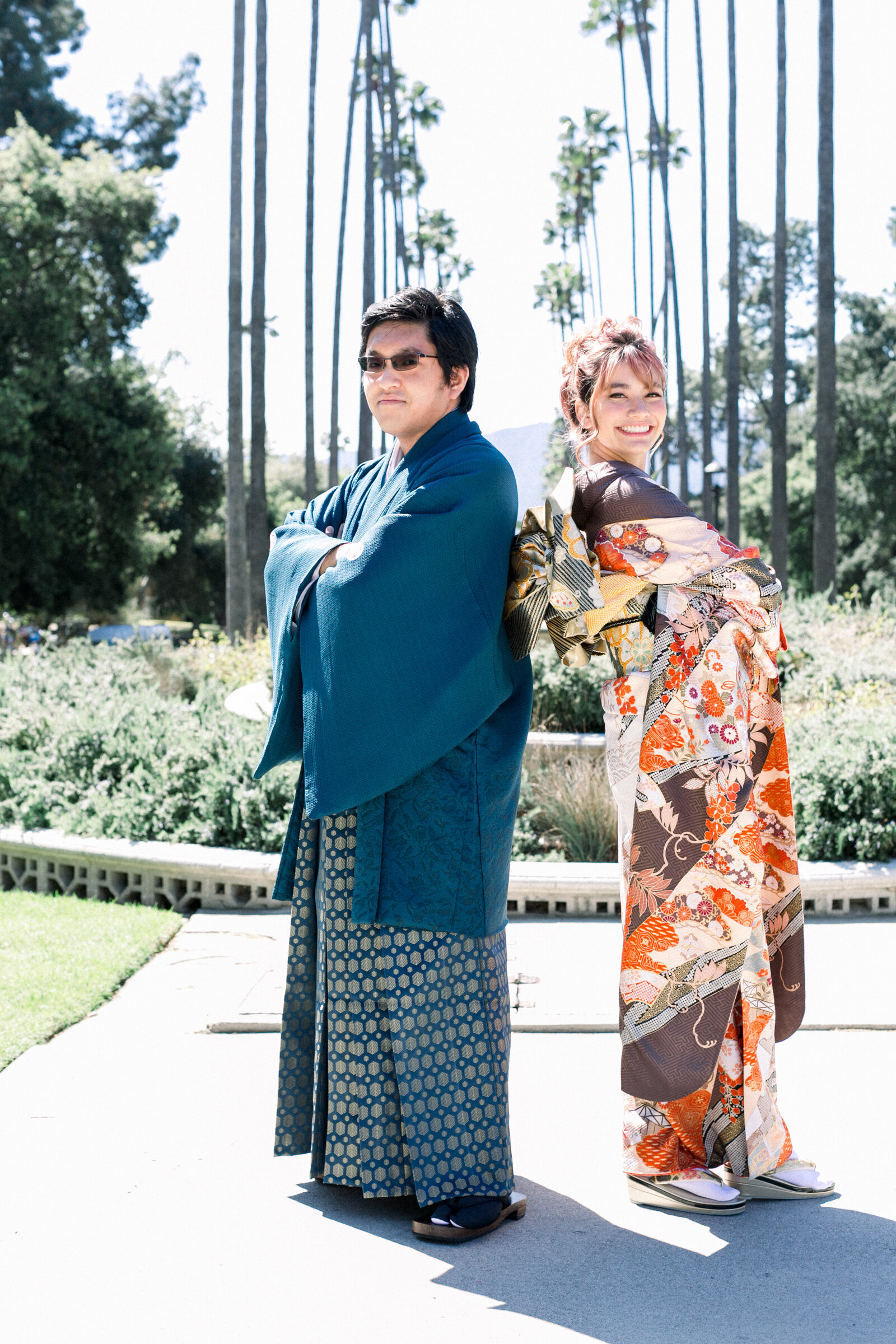 Smiling couple in formal kimono—woman in vibrant furisode and man in deep blue hakama—posing back-to-back at Brand Park for a Seijinshiki (coming-of-age) celebration, kimono rental in Los Angeles | ブランドパークでの成人式。振袖と袴姿のカップルが背中合わせで立つポートレート、着物レンタル。