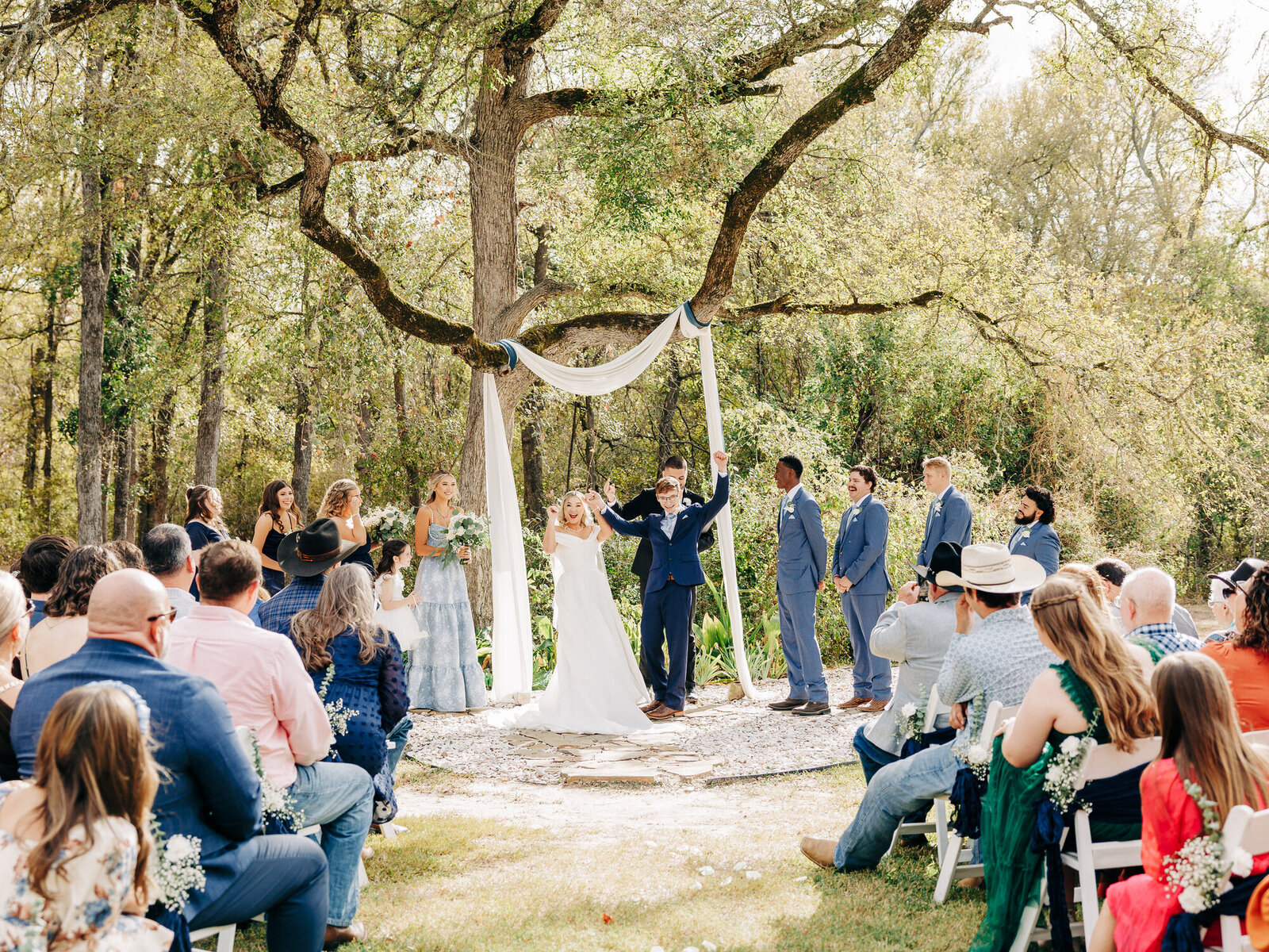 A bride and groom hold hands under a draped arch outdoors, surrounded by their wedding party. Guests sit on white chairs facing them with trees and greenery in the background on a sunny day. The venue is near Austin, TX.