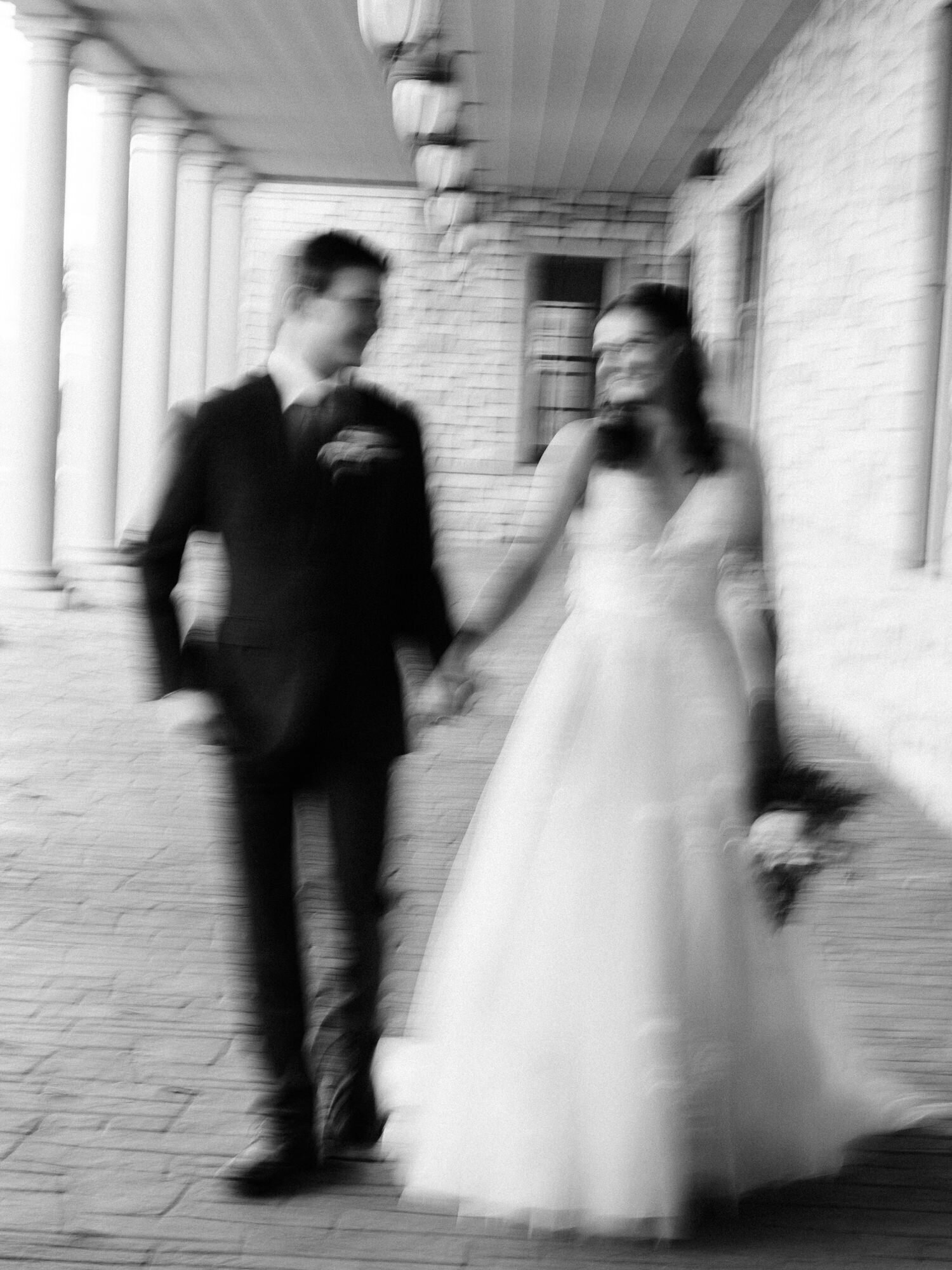 This is an artistic, editorial, film inspired image taken by KD Captures, a wedding photography team in Austin. The image features a bride and a groom walking while looking at each other. The image is black and white, and has a motion blur effect.