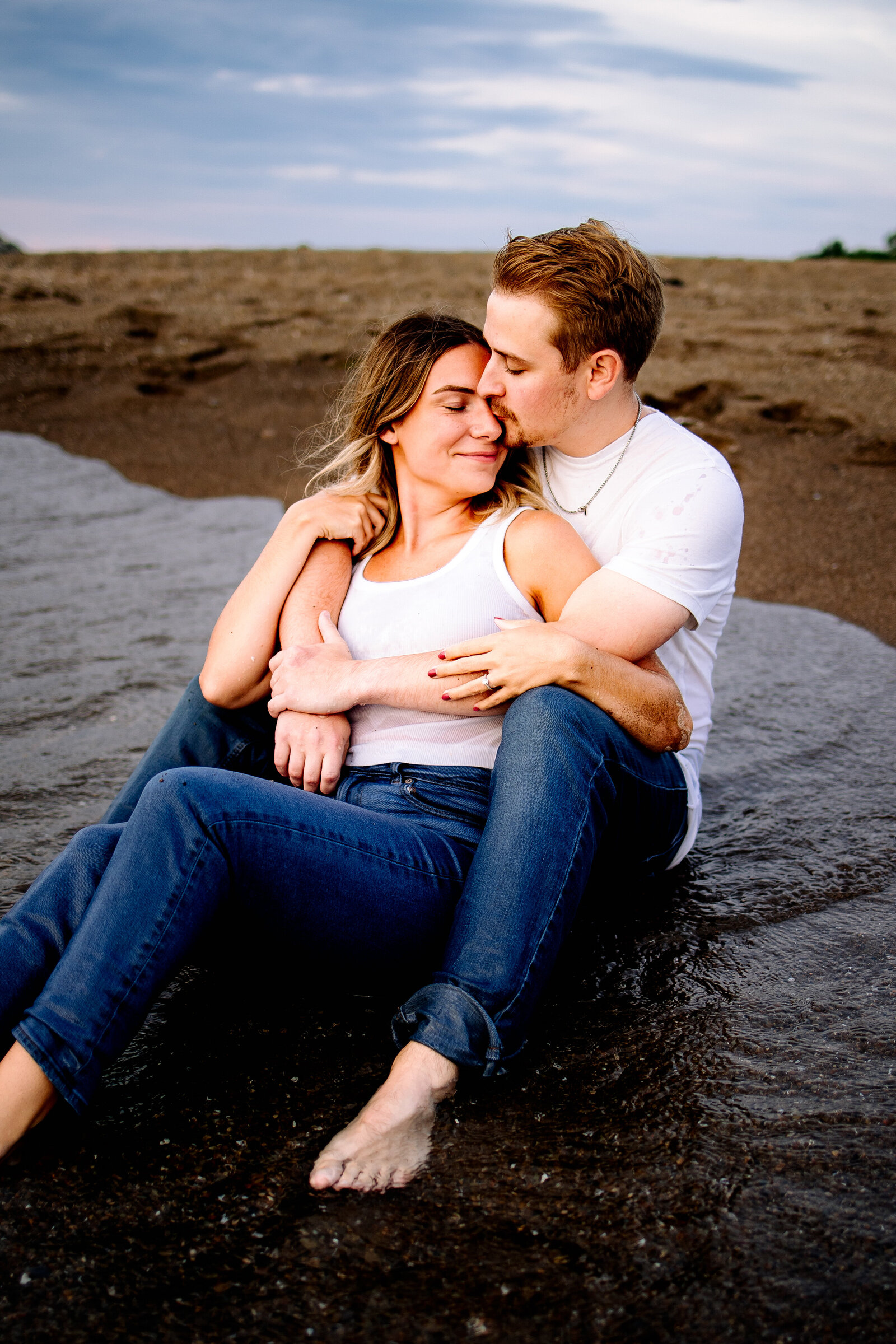Sitting on the waters edge, man wraps up his fiancé from behind and kisses her on the cheek