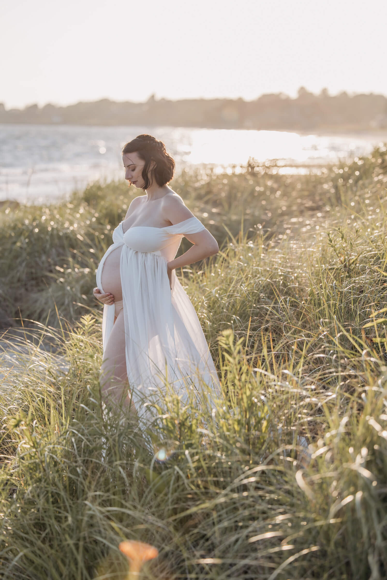 Pregnant woman in white sheer open dress in tall grass in glowy sunlight