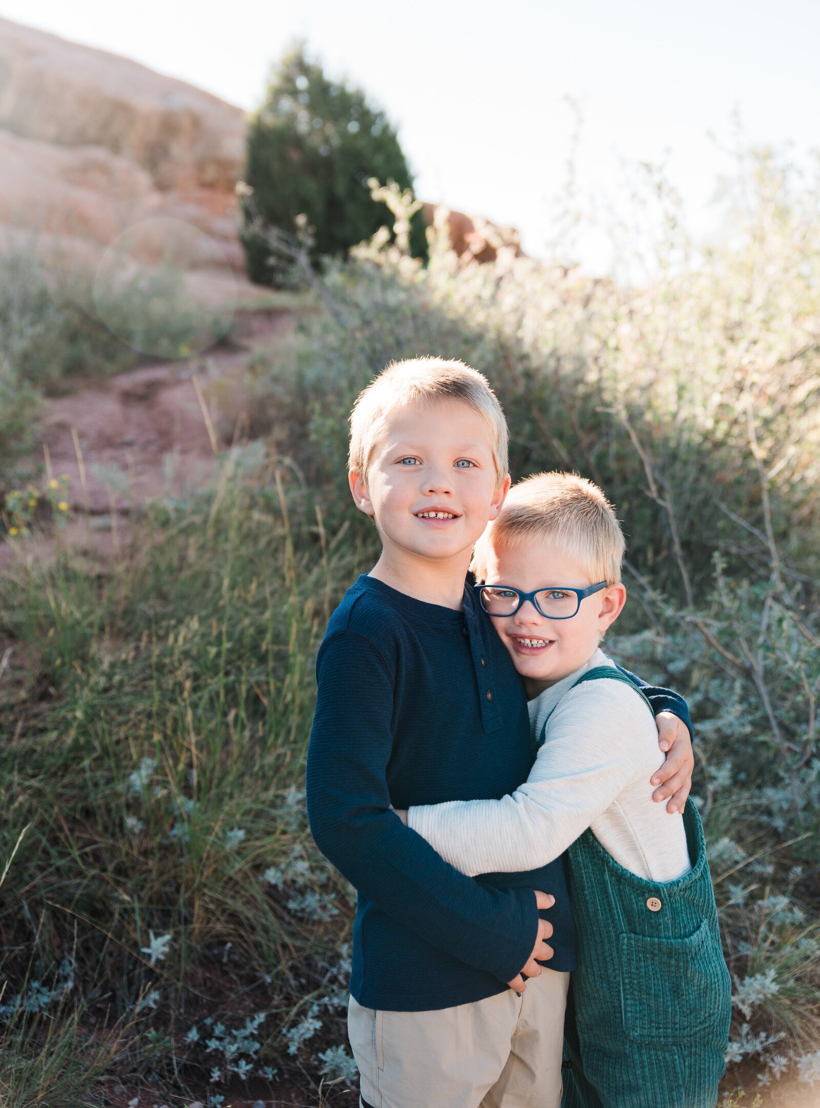 Two young boys hug and smile for denver family photographer outside in front of green shubbery and rocks in the background