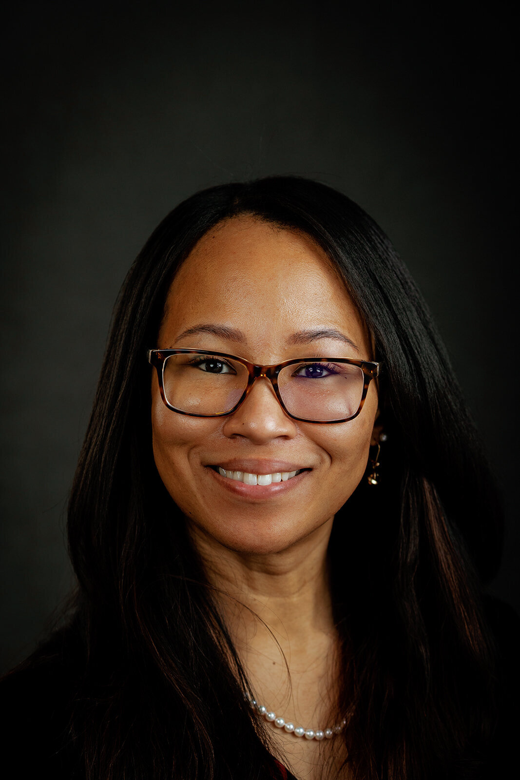 Executive headshot of a woman with glasses, long black hair, and a pearl necklace, smiling warmly against a dark background.