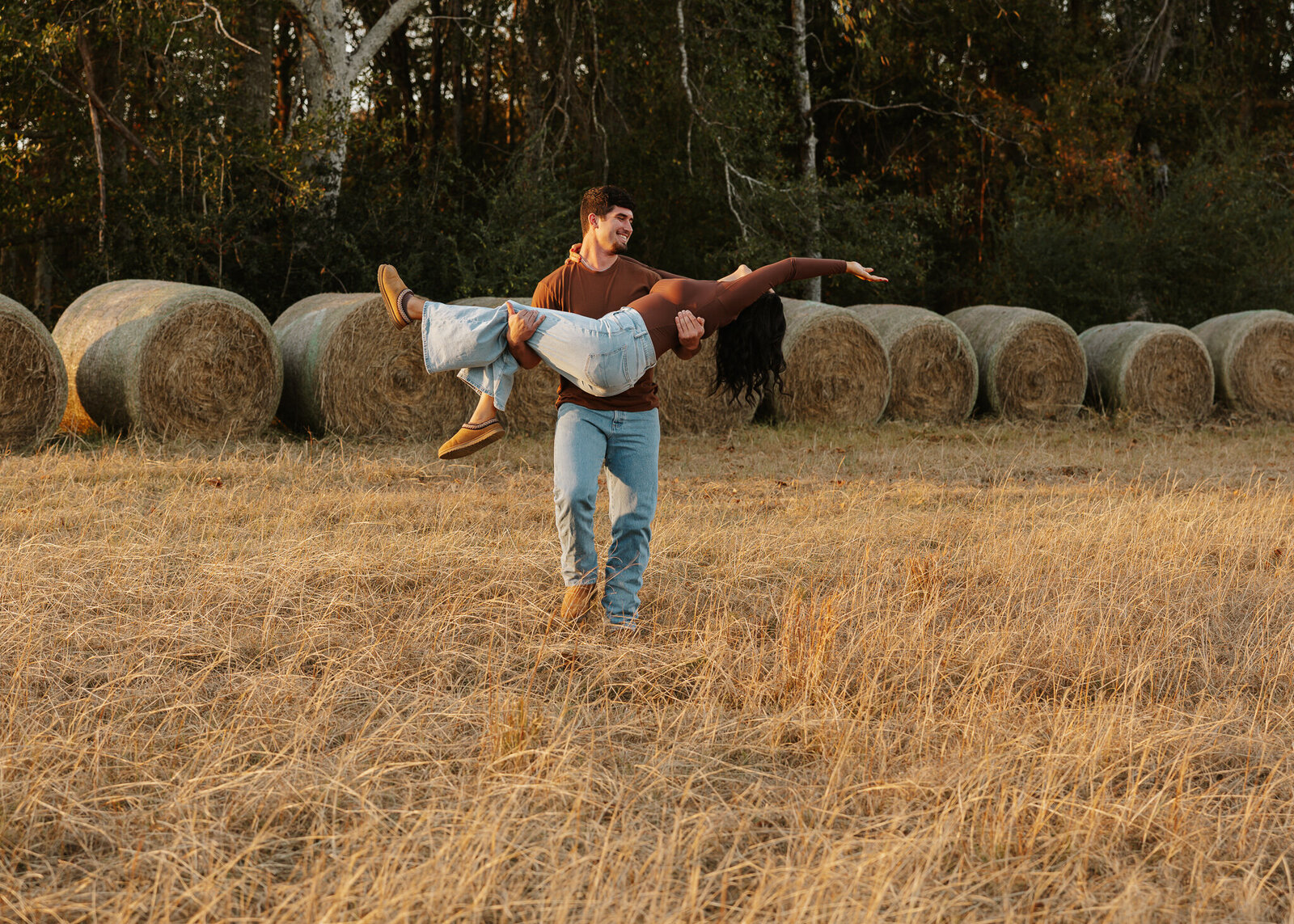 Couples session in a farm field in Aiken SC - boyfriend lifting his girlfriend playfully in front of hay bales during golden hour.