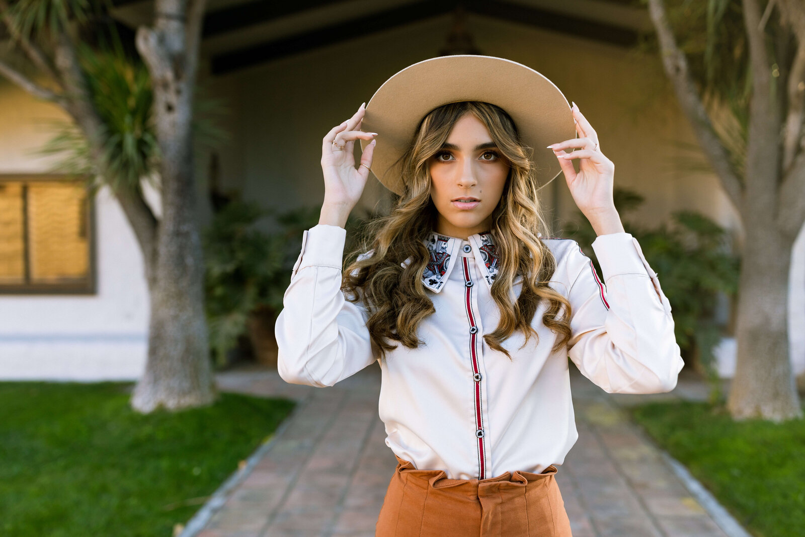 brunette girl wearing brown skirt, white top, and hat standing in front of a house