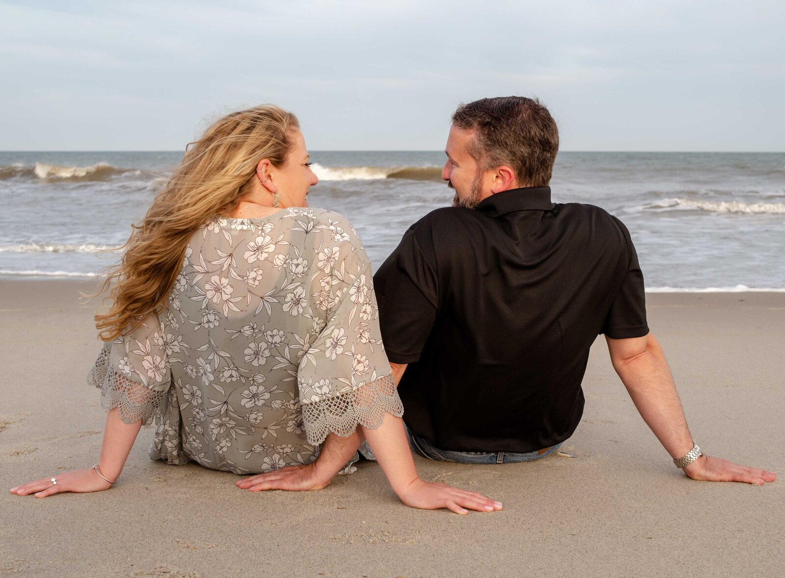 A couple sitting on the sandy beach in Wilmington, NC, gazing at each other with smiles, capturing an authentic moment. This engagement photo showcases the beauty of candid wedding photography in North Carolina, perfect for couples seeking an artistic wedding photographer.