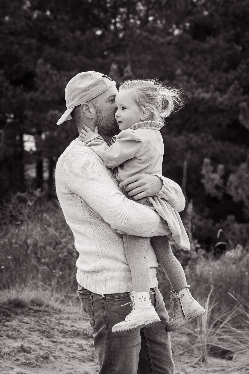 Een dochter wordt door haar vader opgetild en legt haar handen op zijn wangen. Een liefdevol beeld van een fotoshoot in de duinen.