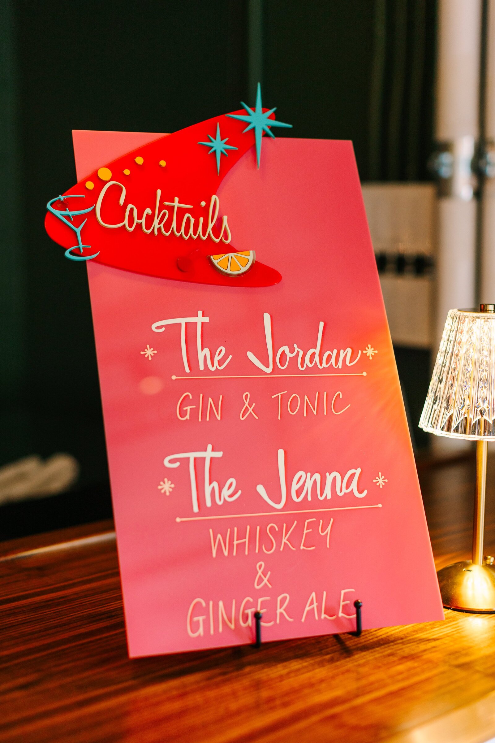 A pink acrylic sign listing signature cocktails named after the bride and groom