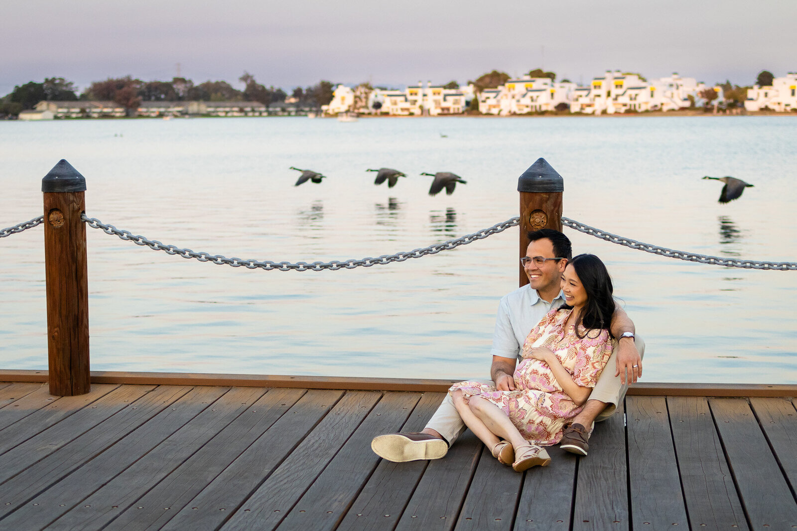 Couple smiles while sitting by the waterfront during a peaceful maternity session at sunset with geese flying overhead — Ellobelle Photography Family Photographer.
