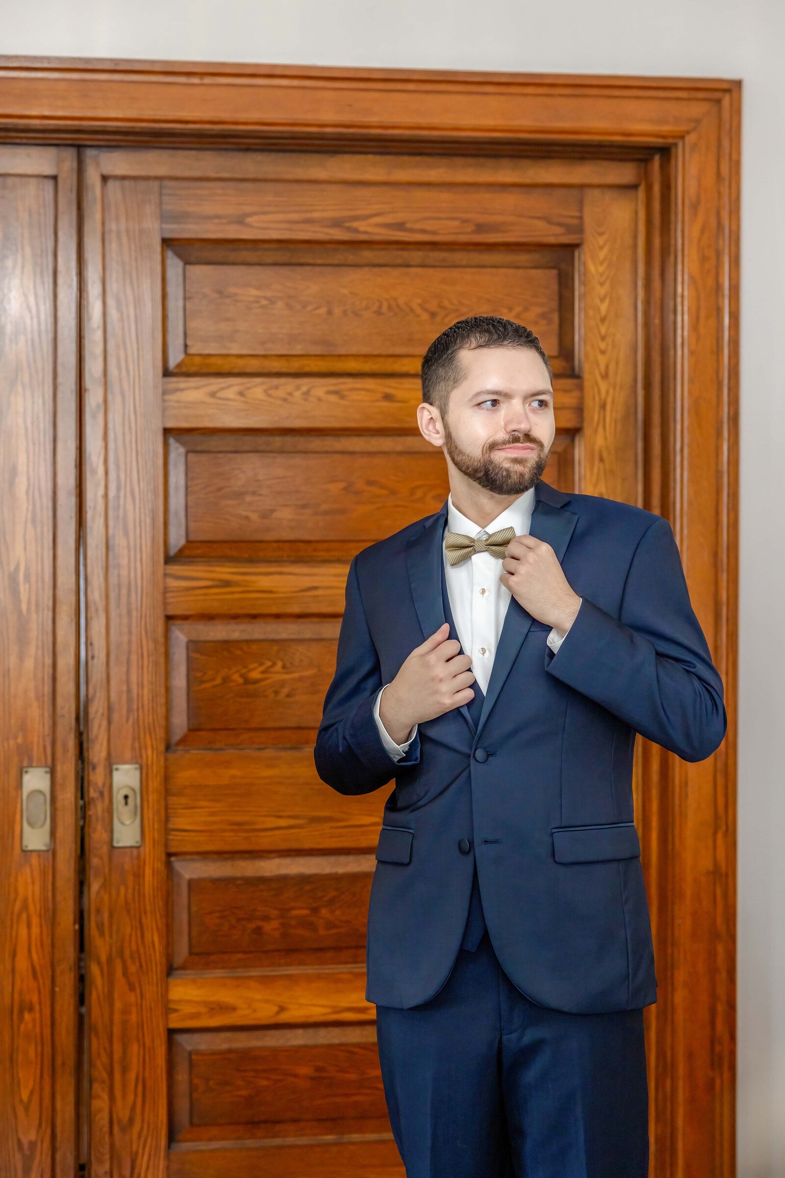 A stylish groom adjusts his bow tie in a navy suit, standing against a wooden door at The Arches wedding venue in Coatesville, PA, blending historic elegance and timeless charm.