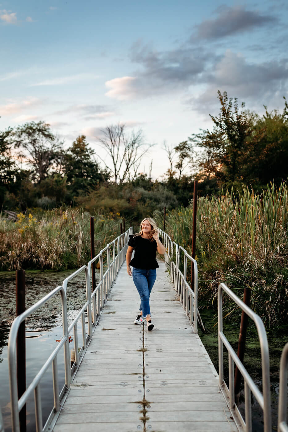 A person walks along a narrow wooden bridge with metal railings, surrounded by tall grasses and trees. The sky is partly cloudy, with hints of sunset colors.