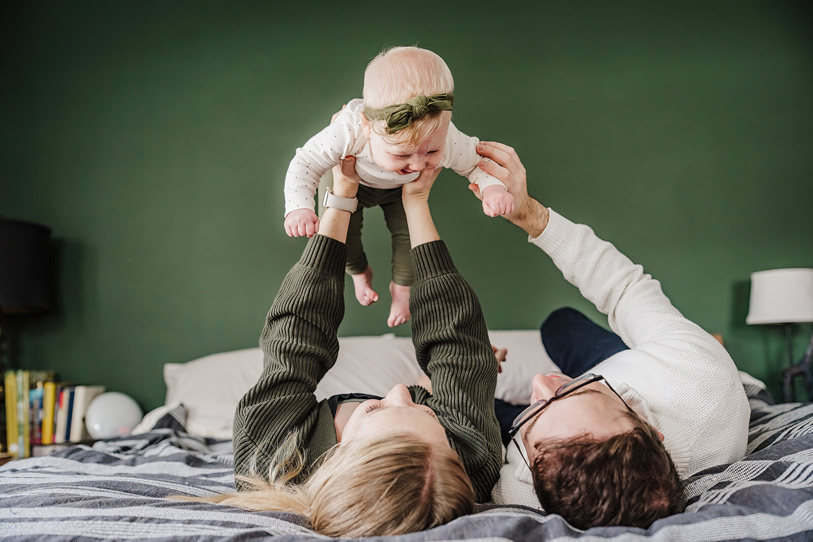 parents hold six month old baby above heads while laying on primary bed