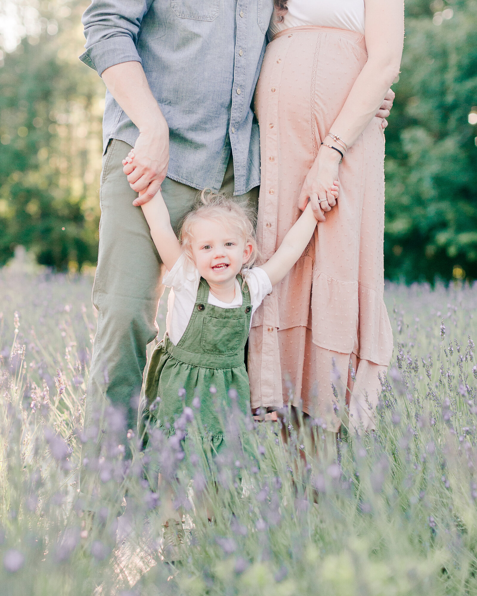 Little girl in green overall dress holding her parents hands in a lavender field by NH newborn photographer Fieldstone Studio.