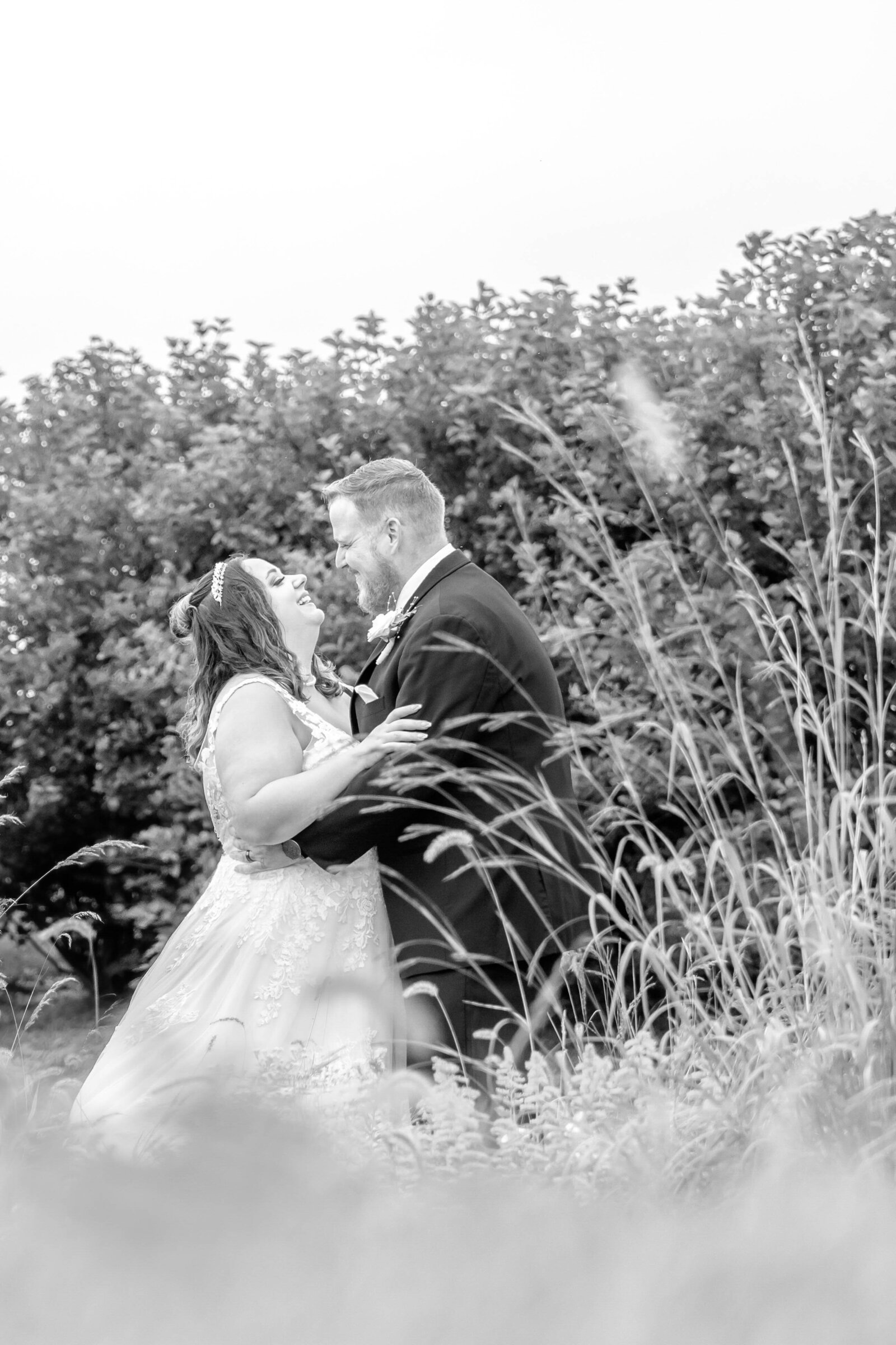 A black-and-white candid of the bride and groom laughing together in a lush meadow surrounded by tall grass, captured during a Asheville, NC wedding, showcasing authentic and romantic moments.