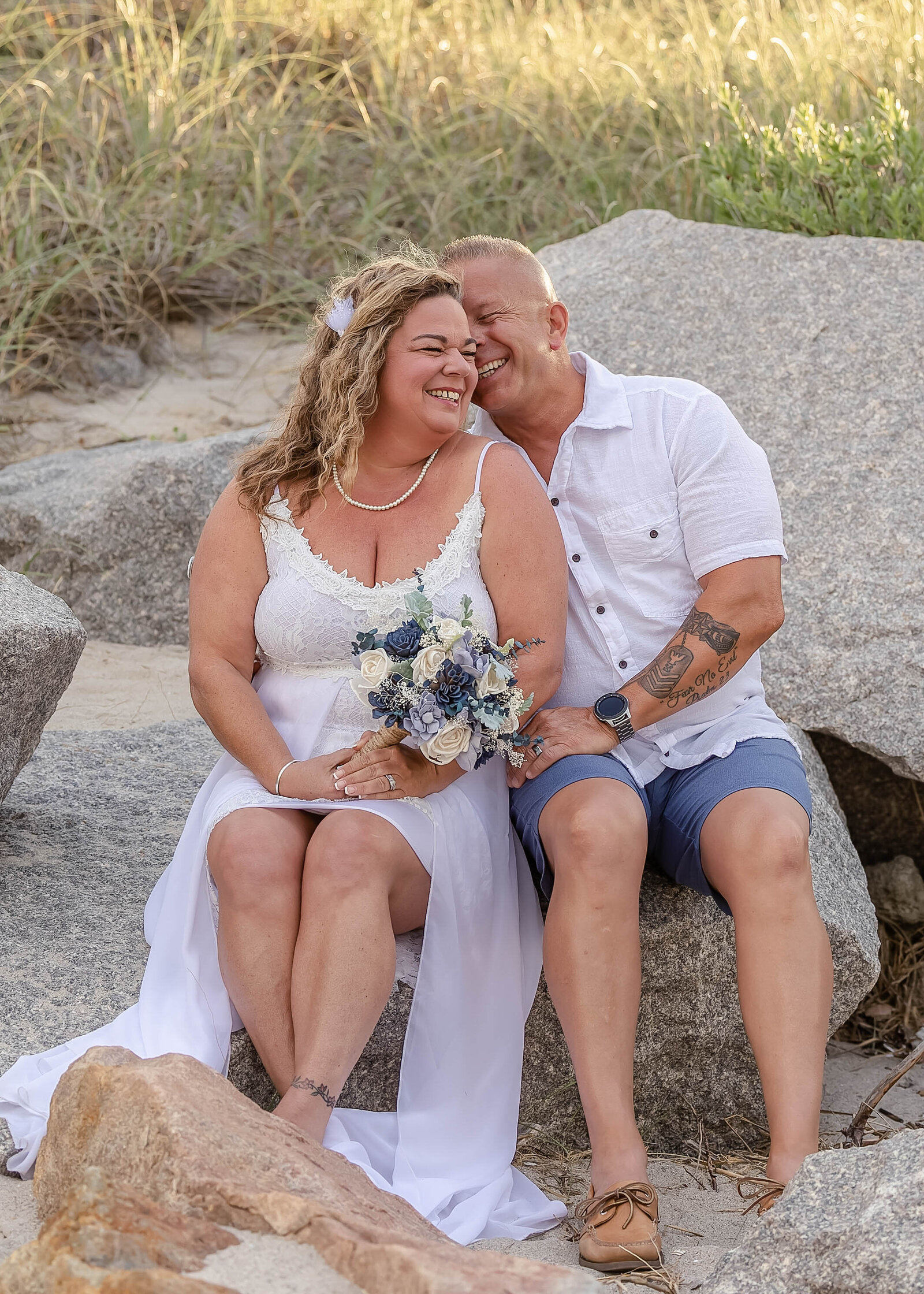The bride and groom share a candid moment sitting on rocks near the beach dunes, with the bride holding a blue and white bouquet, captured during a Kure Beach, NC wedding, blending coastal charm and romance.