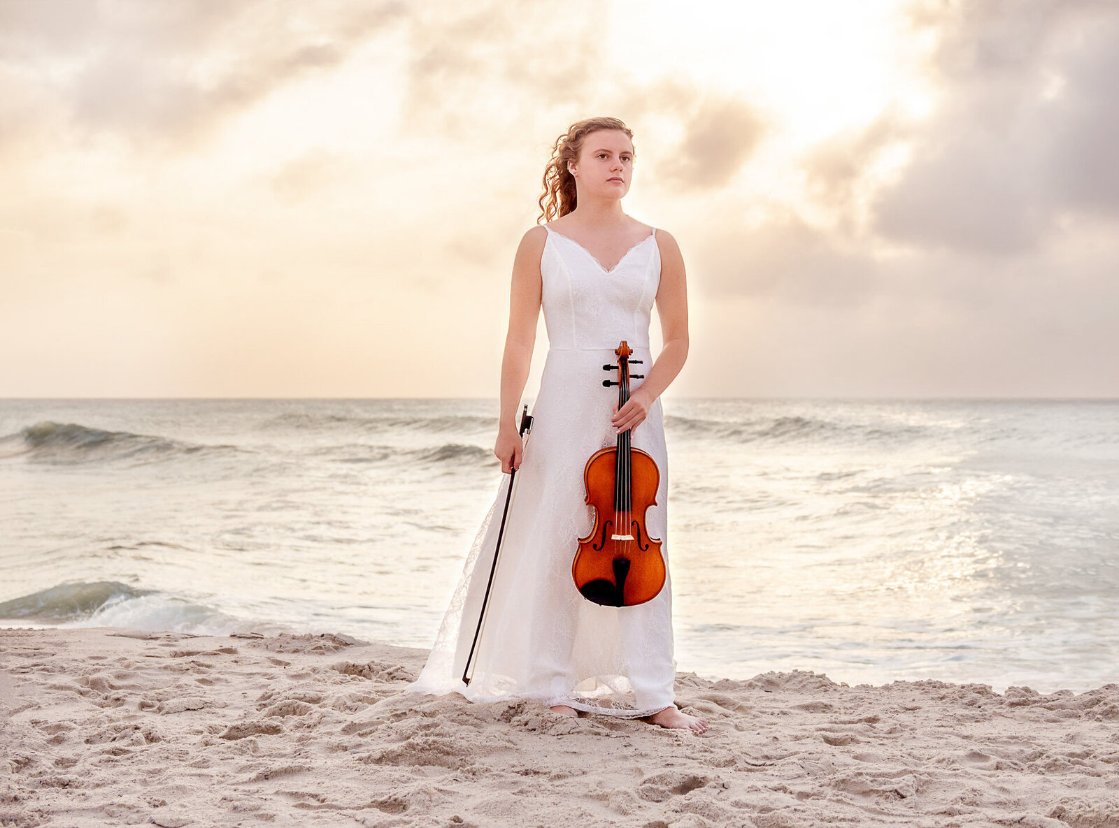 A college senior in a white dress holding a violin, standing on the beach at sunset in North Carolina. This elegant image highlights the blend of musical talent and scenic beauty, perfect for capturing memorable senior photos.