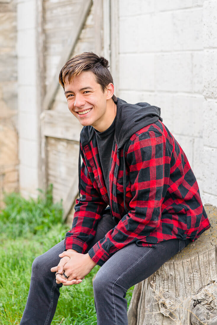 A high school senior boy wearing a red plaid jacket sits on a tree stump in front of a white brick wall in Lehi in the spring. Captured by Salt Lake City senior photographer Melissa Woodruff Photography