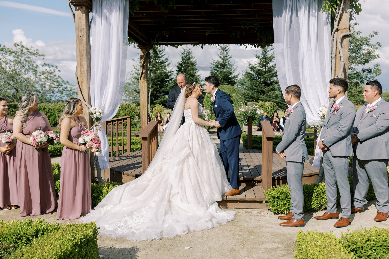 outdoor ceremony for a Bay Area wedding with bride and groom holding hands and kissing one another while their wedding party stands around them in a pergola captured by sacramento wedding photographer