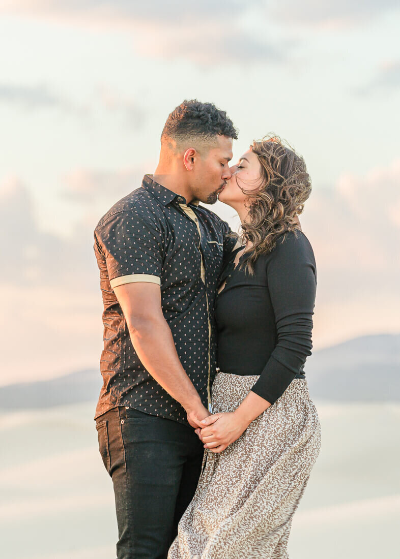 An engaged couple kissing and holding hands while standing on a sand dune at Little Sahara, Nephi. Captured by Utah Engagement Photographer Melissa Woodruff Photography