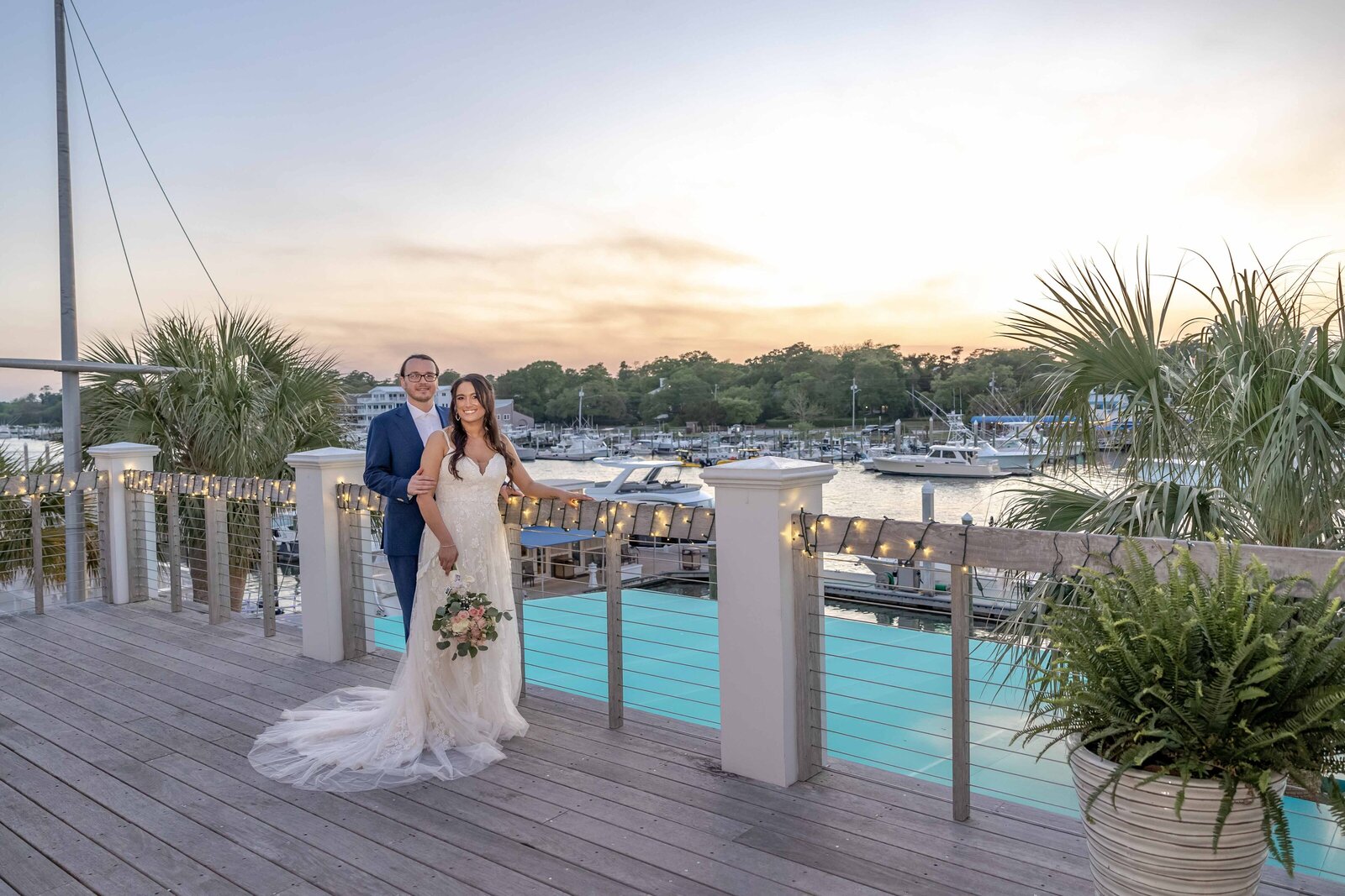 The bride and groom pose on a marina dock at sunset, surrounded by string lights, palm trees, and yachts, captured during a Wrightsville Beach, NC wedding, showcasing coastal elegance and romantic charm.