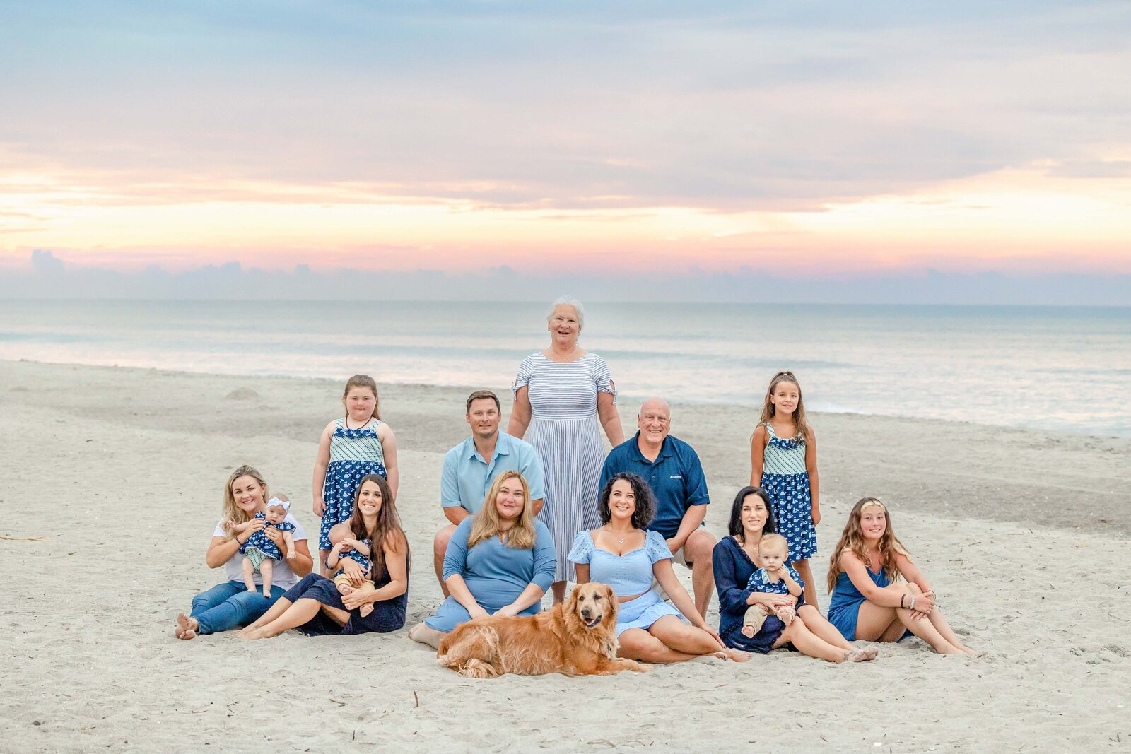 A large family, including a golden retriever, sits on the beach at sunset, dressed in blue and white outfits. The pastel sky adds to the serene atmosphere, perfect for artistic family photography in Wilmington, NC, capturing peaceful beachside gatherings.